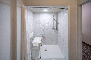 Ducha a pie con pared de azulejos blancos, cabezal de ducha, ducha de mano y una toalla blanca sobre un banco.