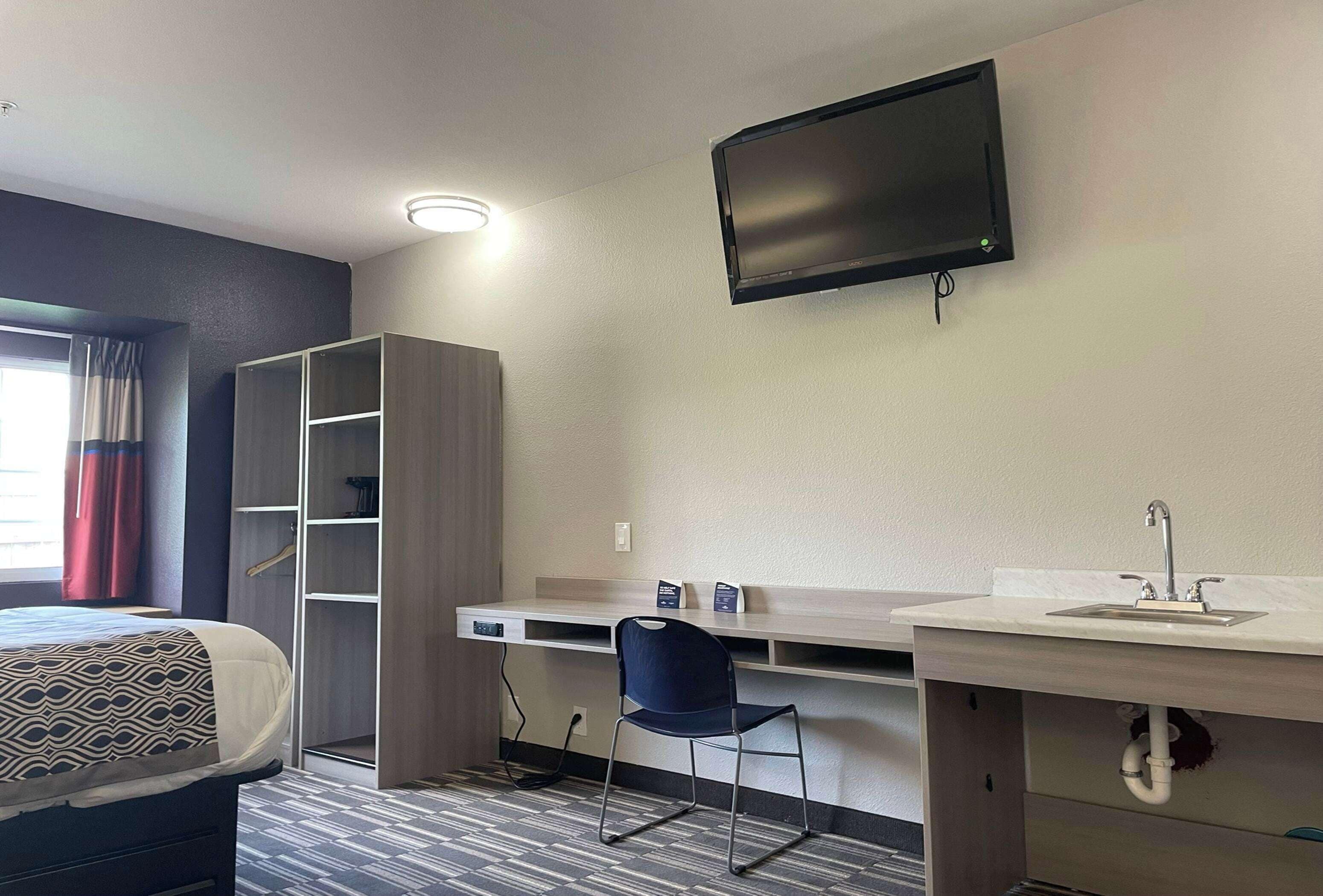 A hotel room with a bed, a desk with a chair, a TV mounted on the wall, a built-in shelf, and a sink.