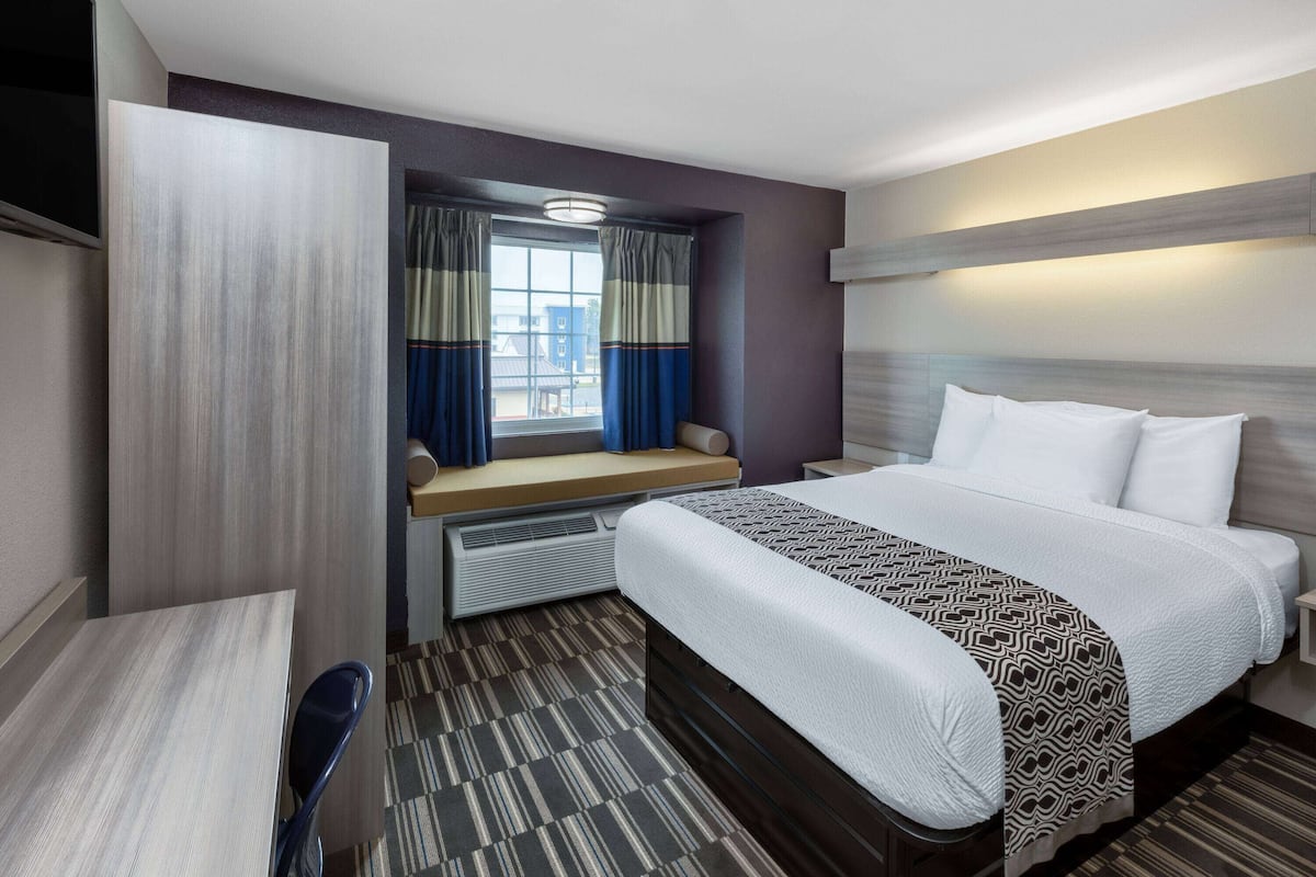 Une chambre d’hôtel équipée d’un lit, d’un bureau, d’une chaise, d’une fenêtre avec des rideaux et d’un téléviseur fixé au mur.