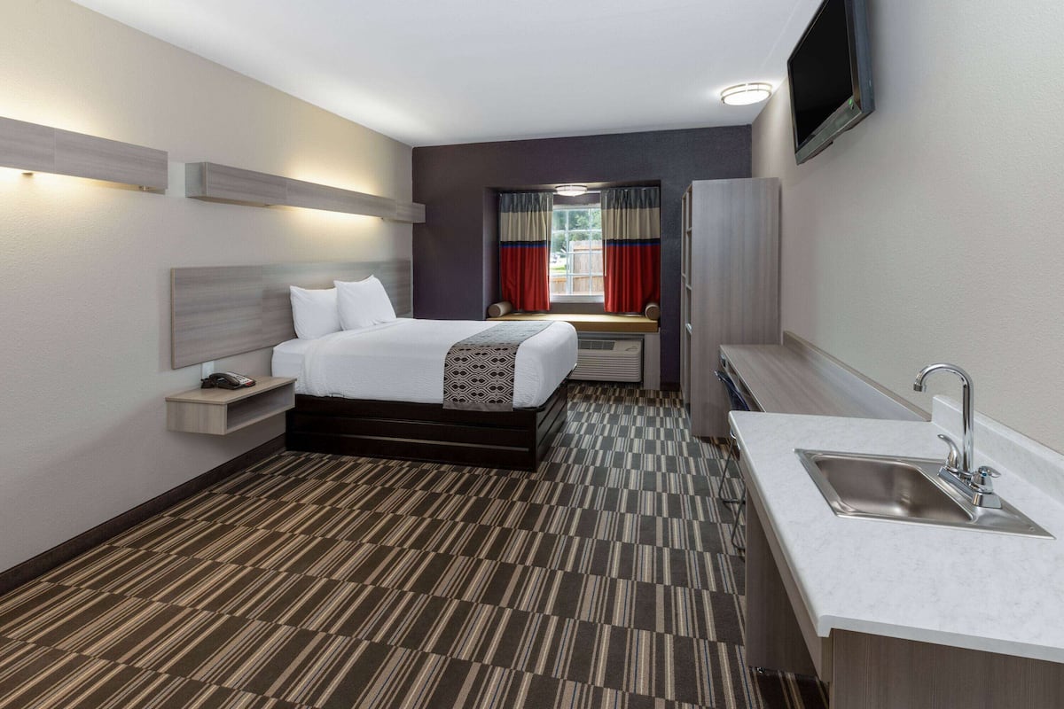 Une chambre d’hôtel comprenant un lit, une table de chevet, une lampe, une fenêtre avec des rideaux et un lavabo.