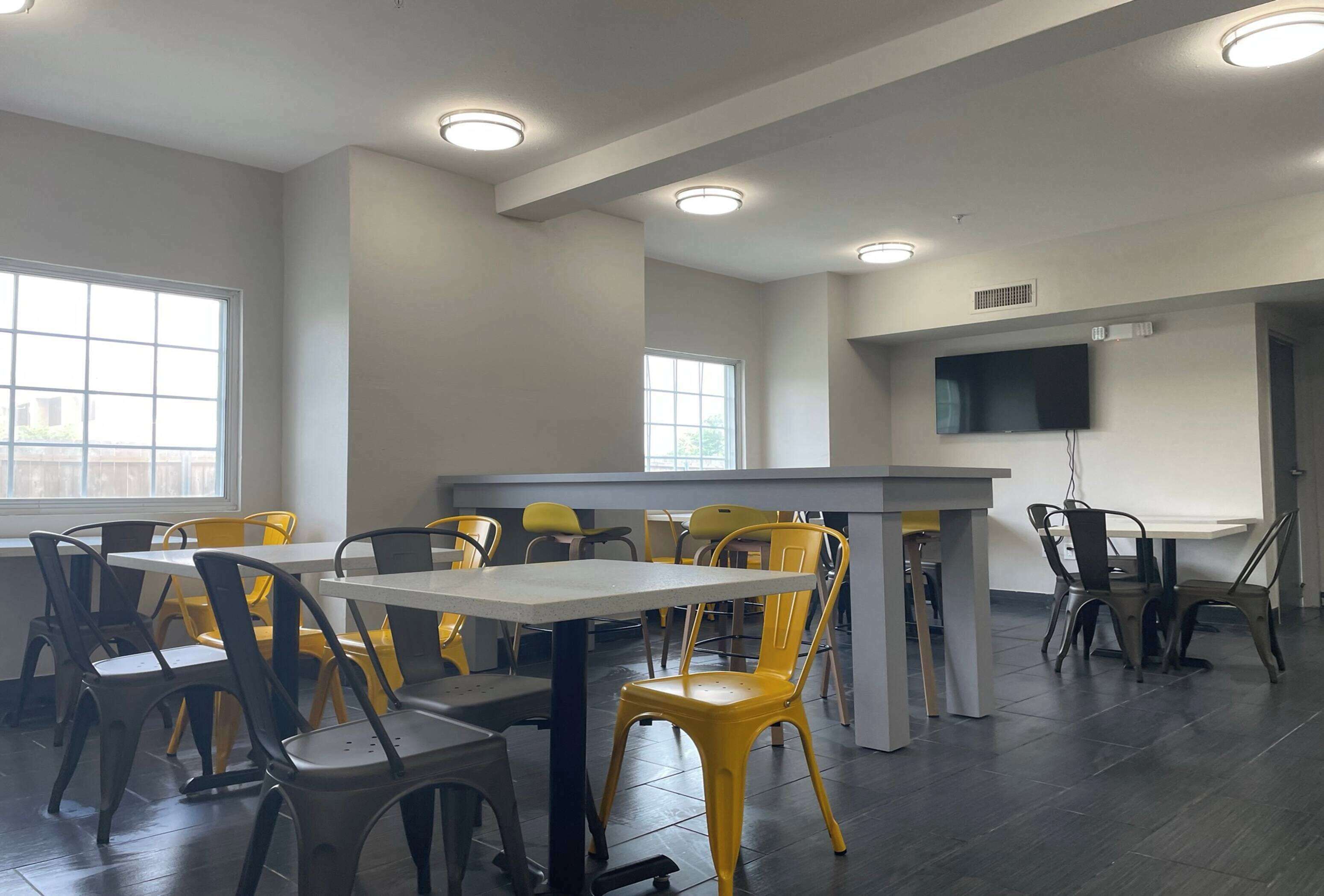 A dining area with tables and chairs, a flat-screen TV mounted on the wall, and large windows providing natural light.