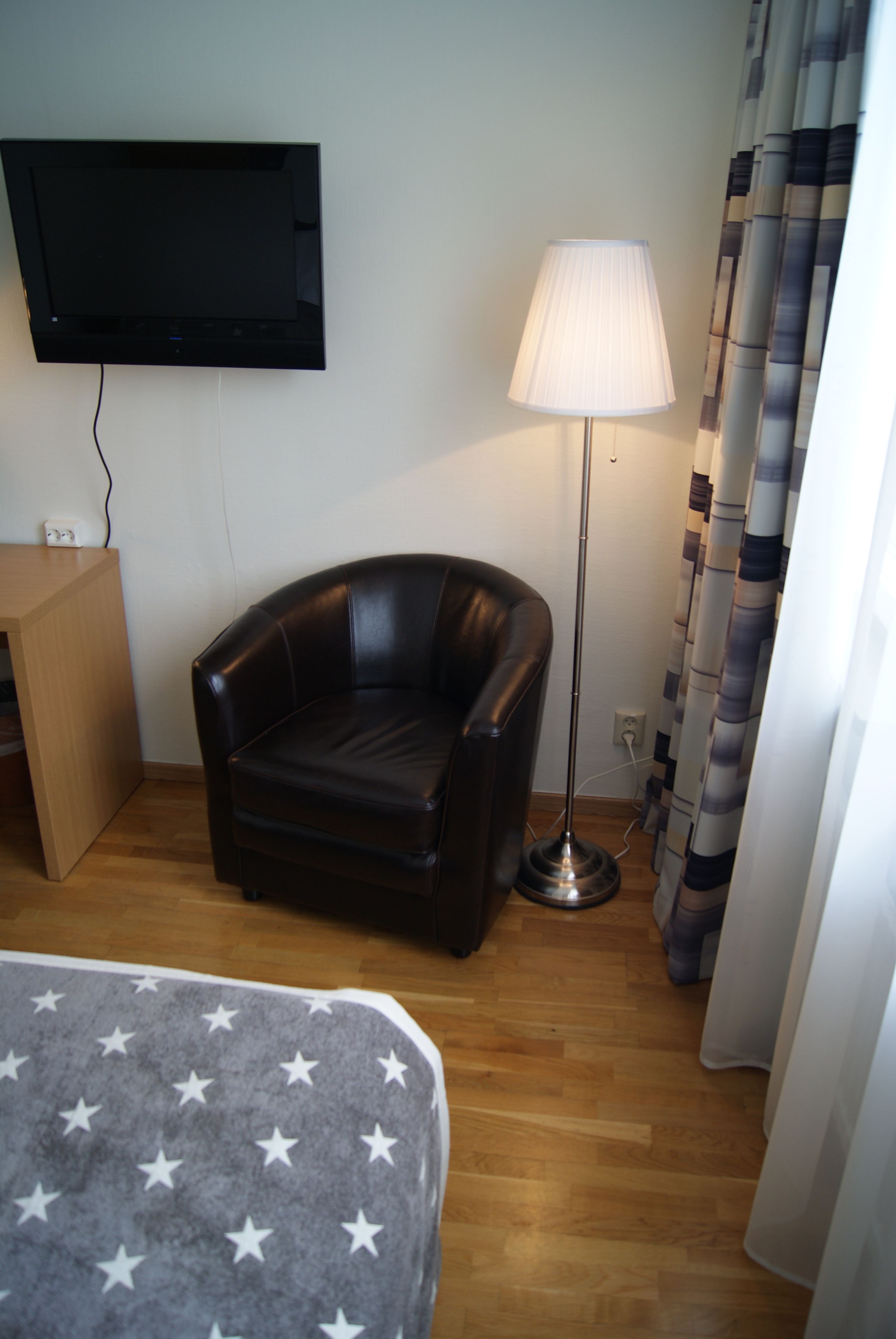 Ett hotellrum med en platt-TV som är monterad på väggen, en svart läderfåtölj, en golvlampa, ett litet träbord och en säng med ett grått överkast prytt med vita stjärnor.