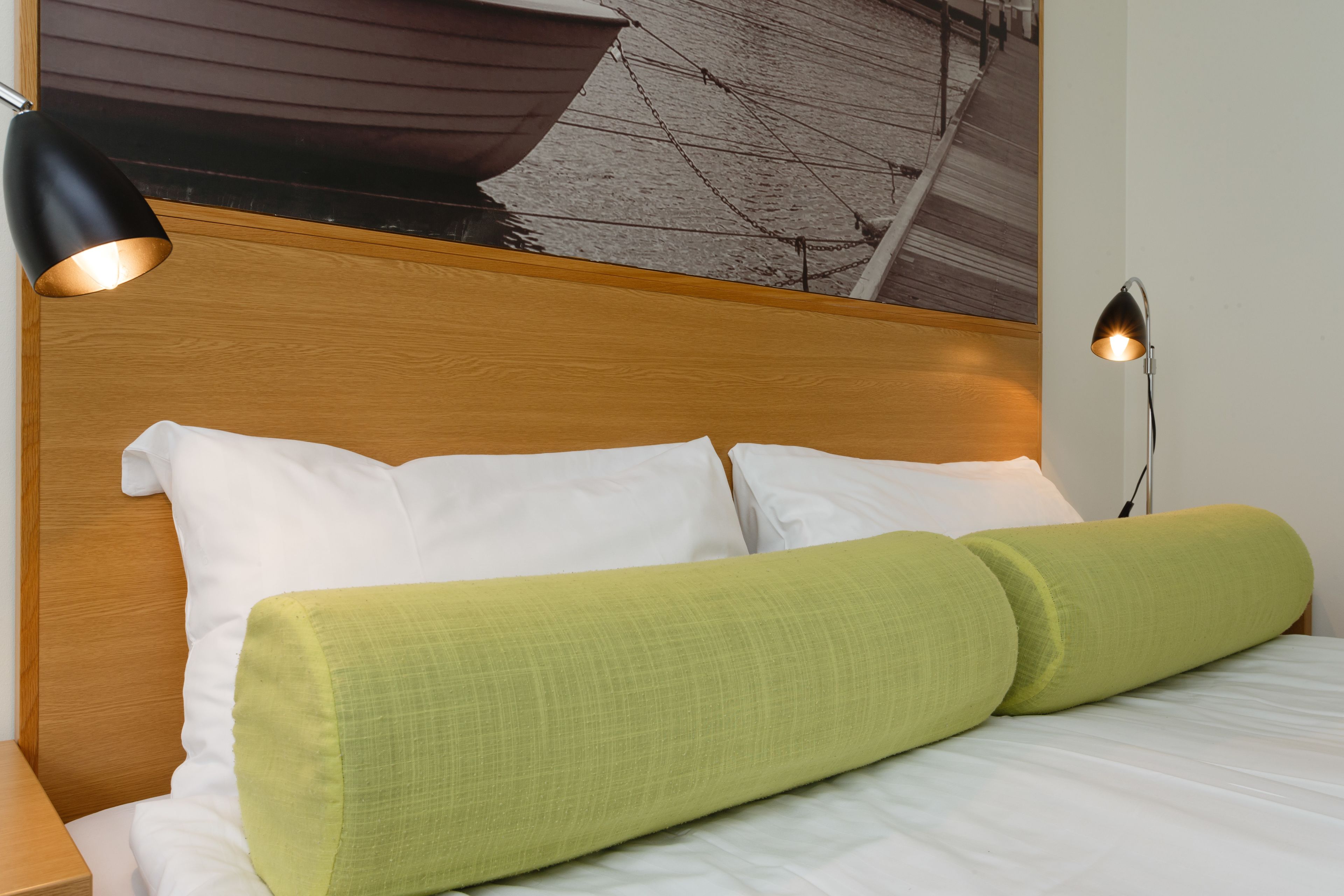 A neatly made bed with white and green bedding, a wooden headboard, and a wall-mounted lamp.