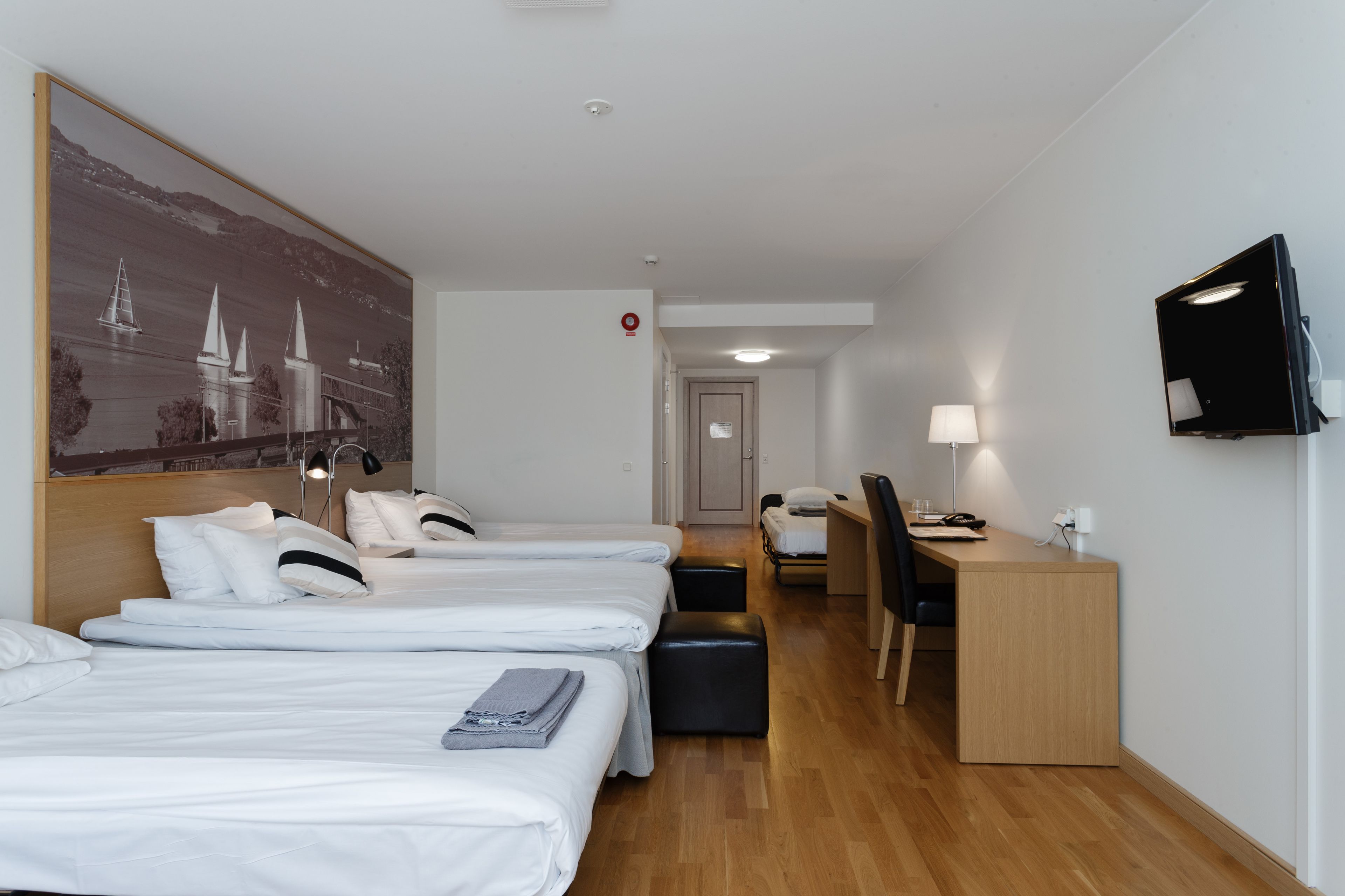 Et hotelværelse med tre senge, et skrivebord, en stol, et fjernsyn og et billede på væggen.