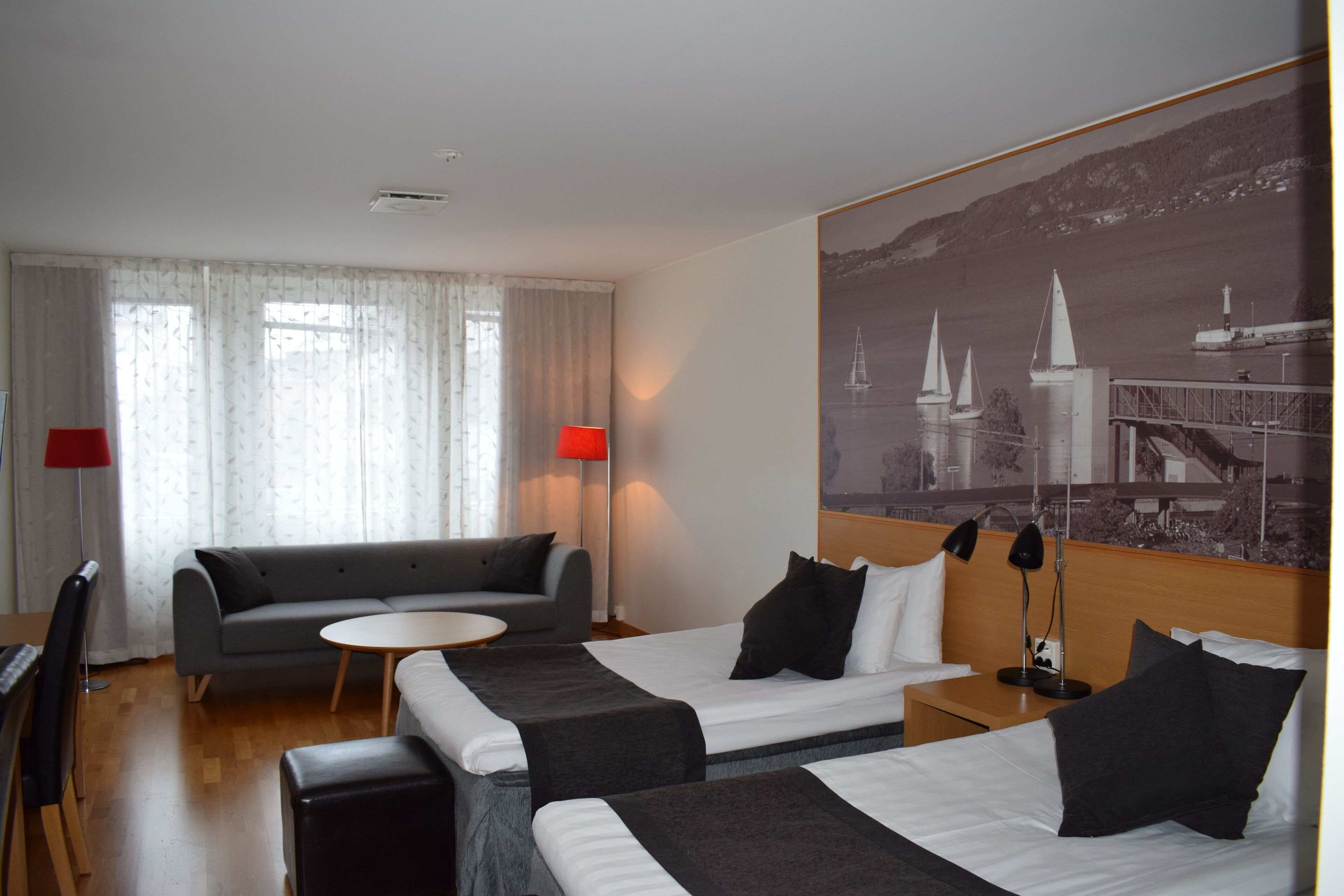 A hotel room with two beds, a sofa, a small round table, and a large window with sheer curtains.