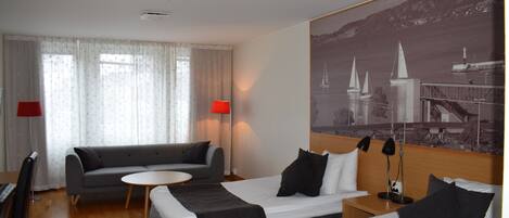 A hotel room with two beds, a sofa, a small round table, and a large window with sheer curtains.