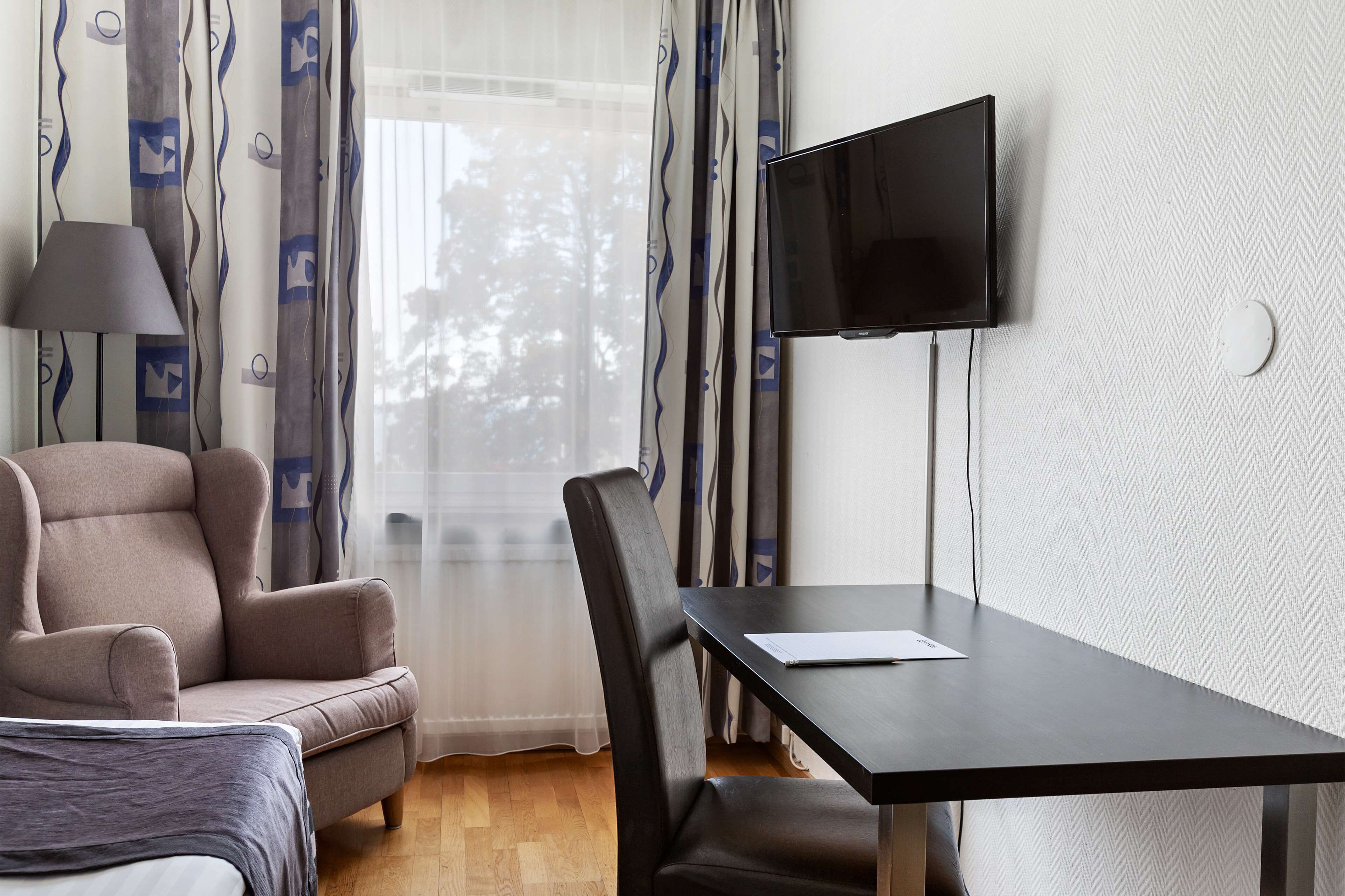 Et hotelværelse med seng, sofa, spisebord, tv ophængt på væggen og et vindue med gardiner.
