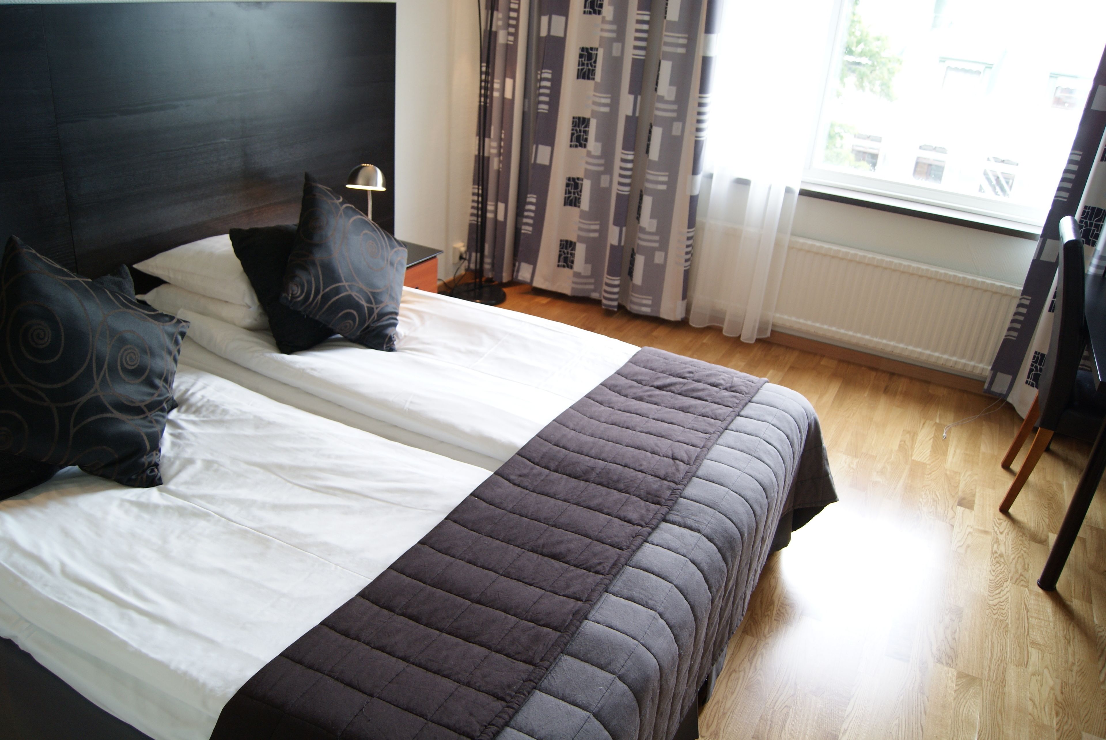 A double bed with a dark quilt and white linens, a bedside table with a lamp, patterned curtains, and a view of the outdoors through a window.