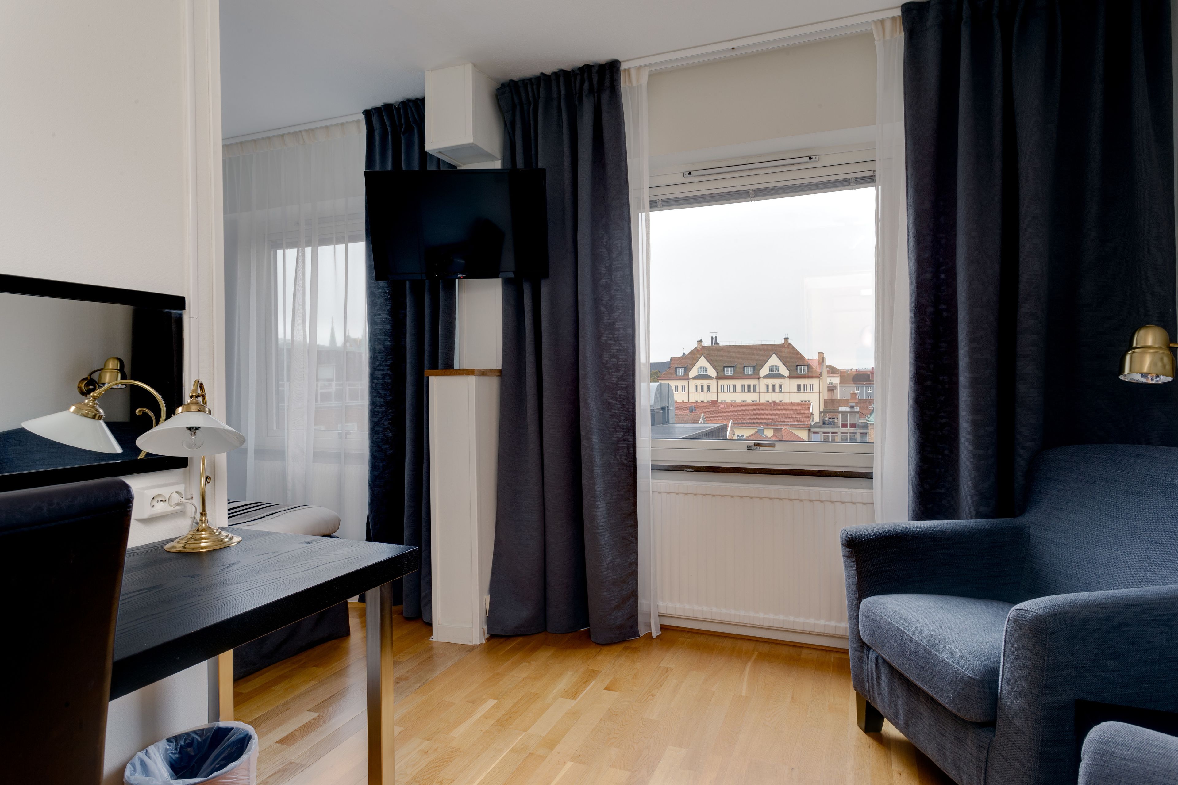 Una habitación de hotel moderna con suelo de madera, un escritorio negro con lámpara, un televisor montado en la pared y vistas a edificios a través de la ventana.