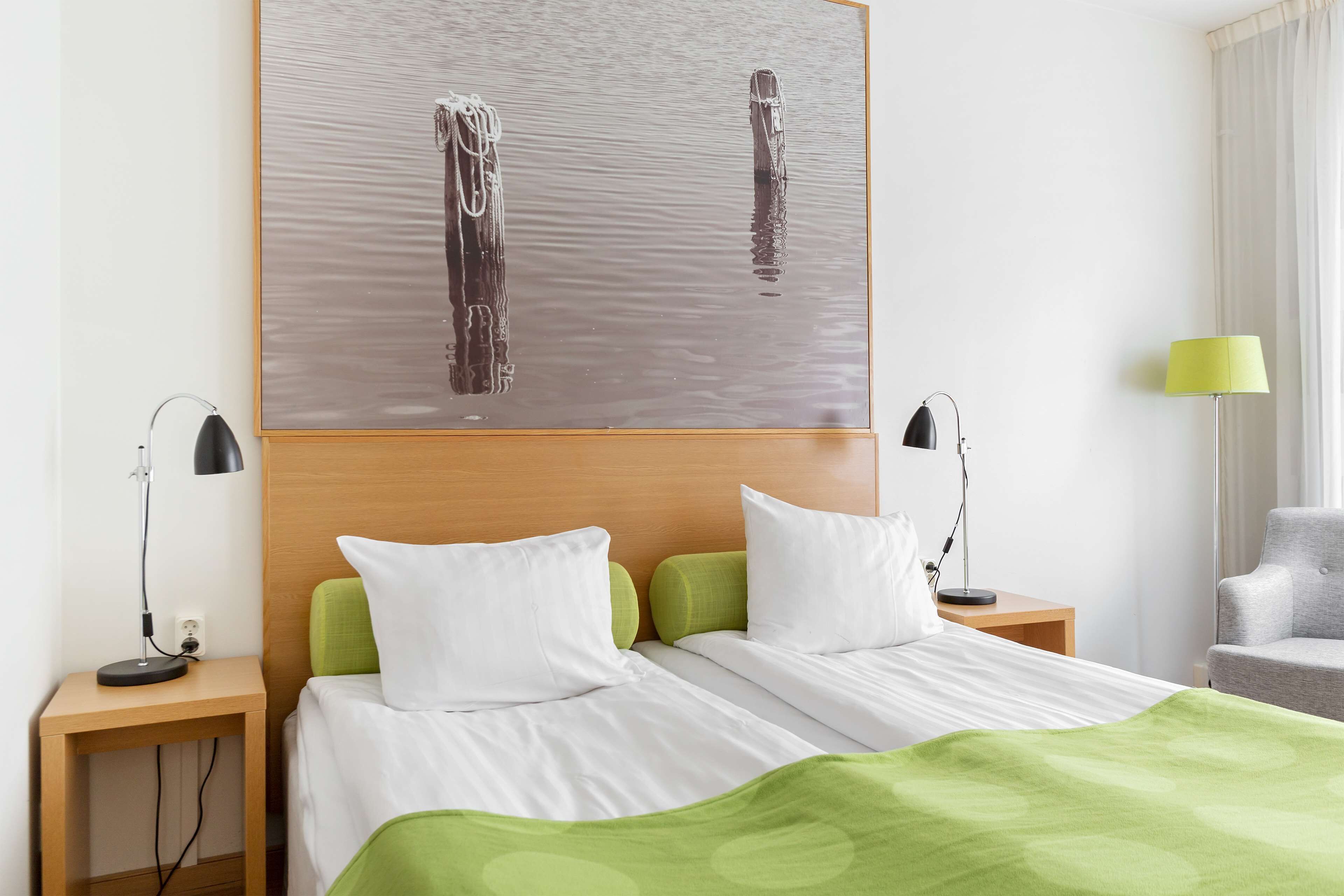 A hotel room with a bed, two bedside tables with lamps, a green blanket, and a painting of two figures in water.