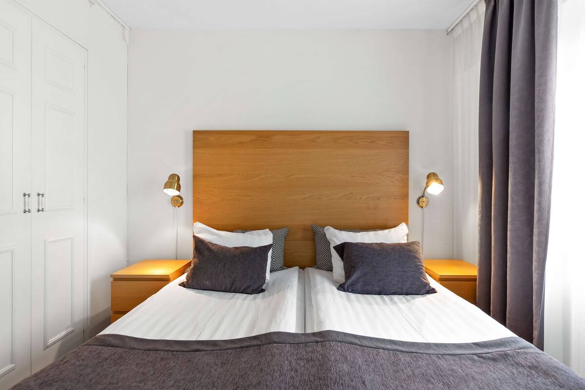 A double bed with a wooden headboard, two bedside tables with lamps, and a large window with curtains.