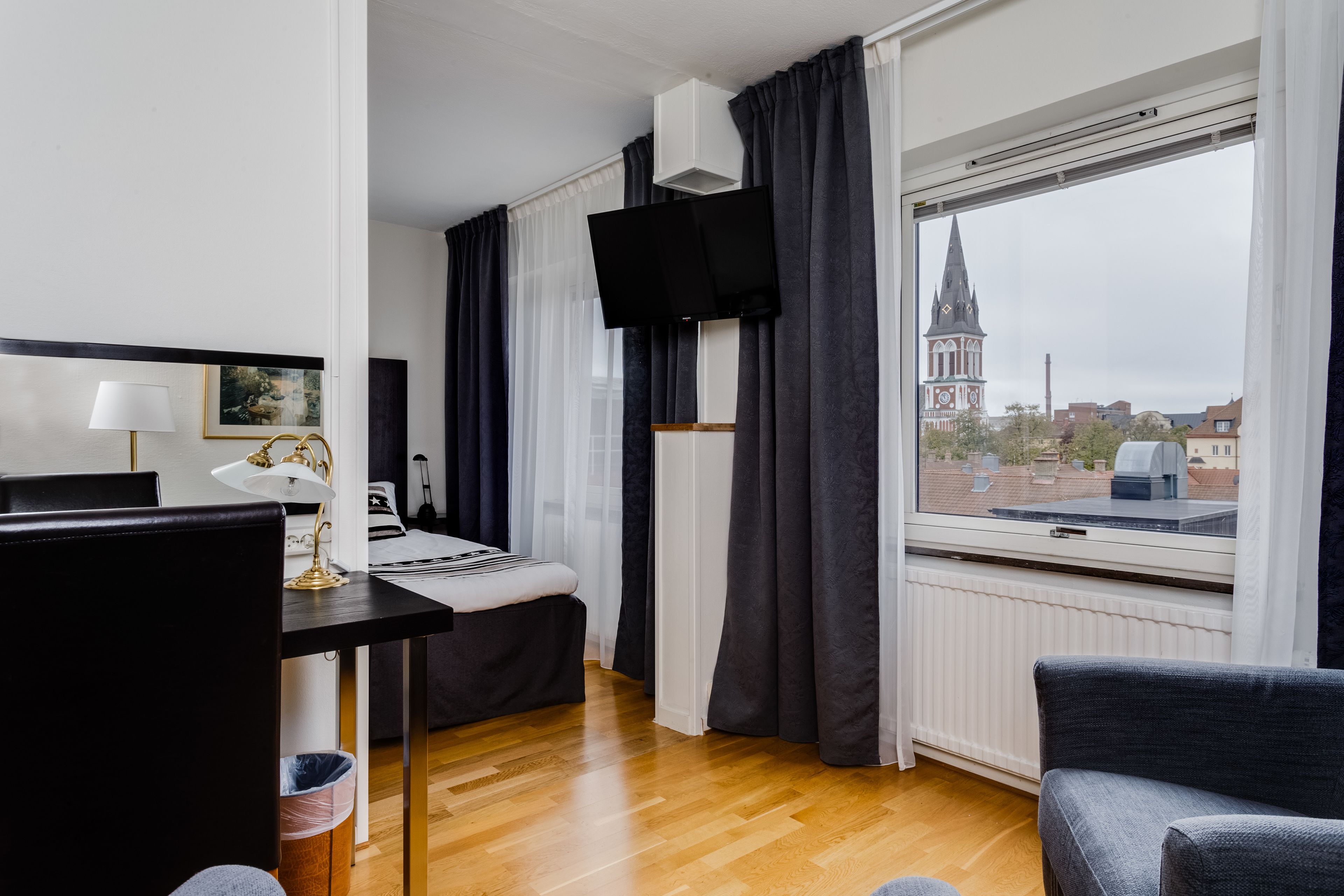 A hotel room with a bed, a desk with a lamp, a TV mounted on the wall, and a view of a church spire through the window.