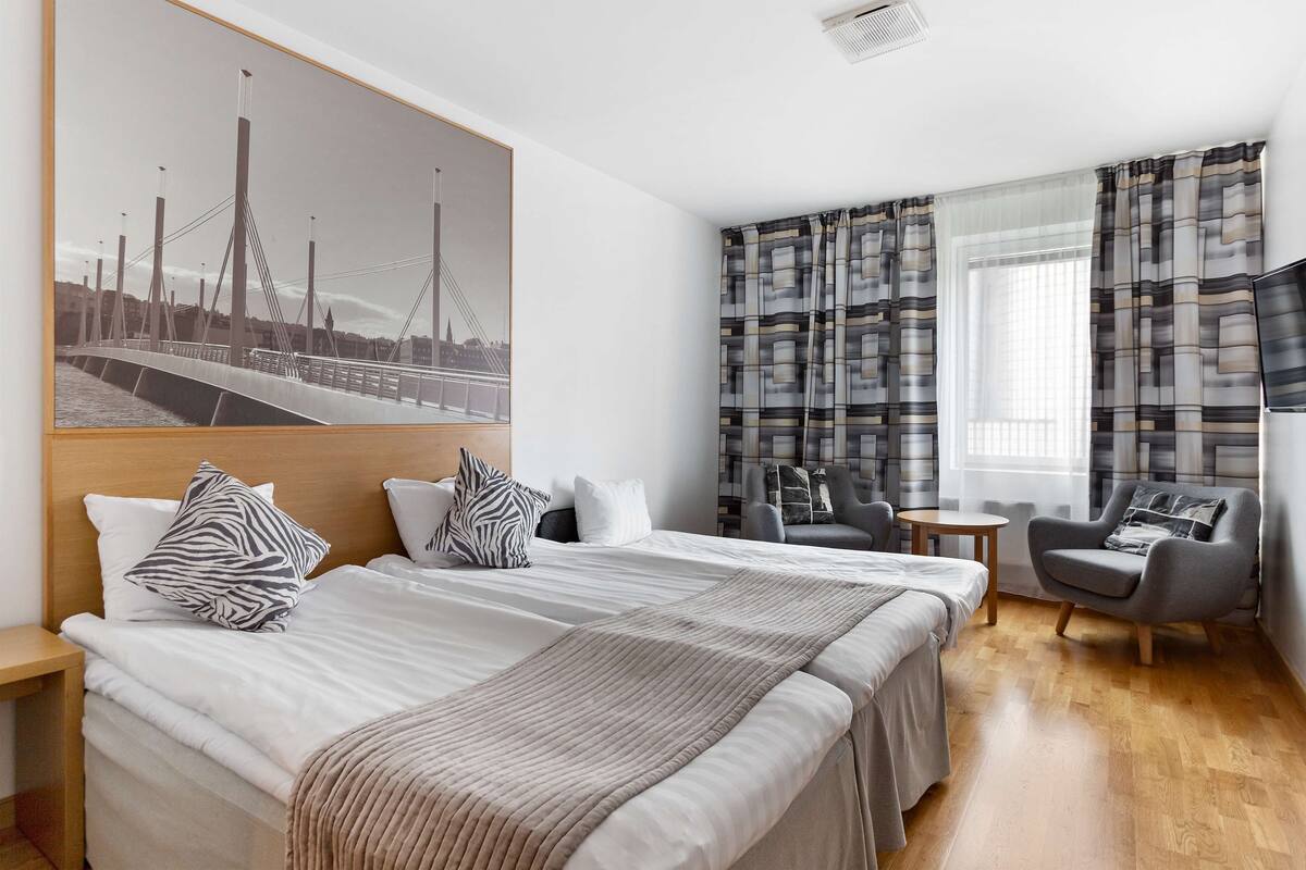A hotel room with a large bed, a desk, a chair, a TV, and a window with curtains.