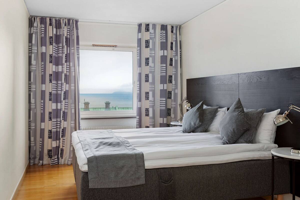 A modern bedroom with a large bed, grey bedding, and a view of the sea through a window.