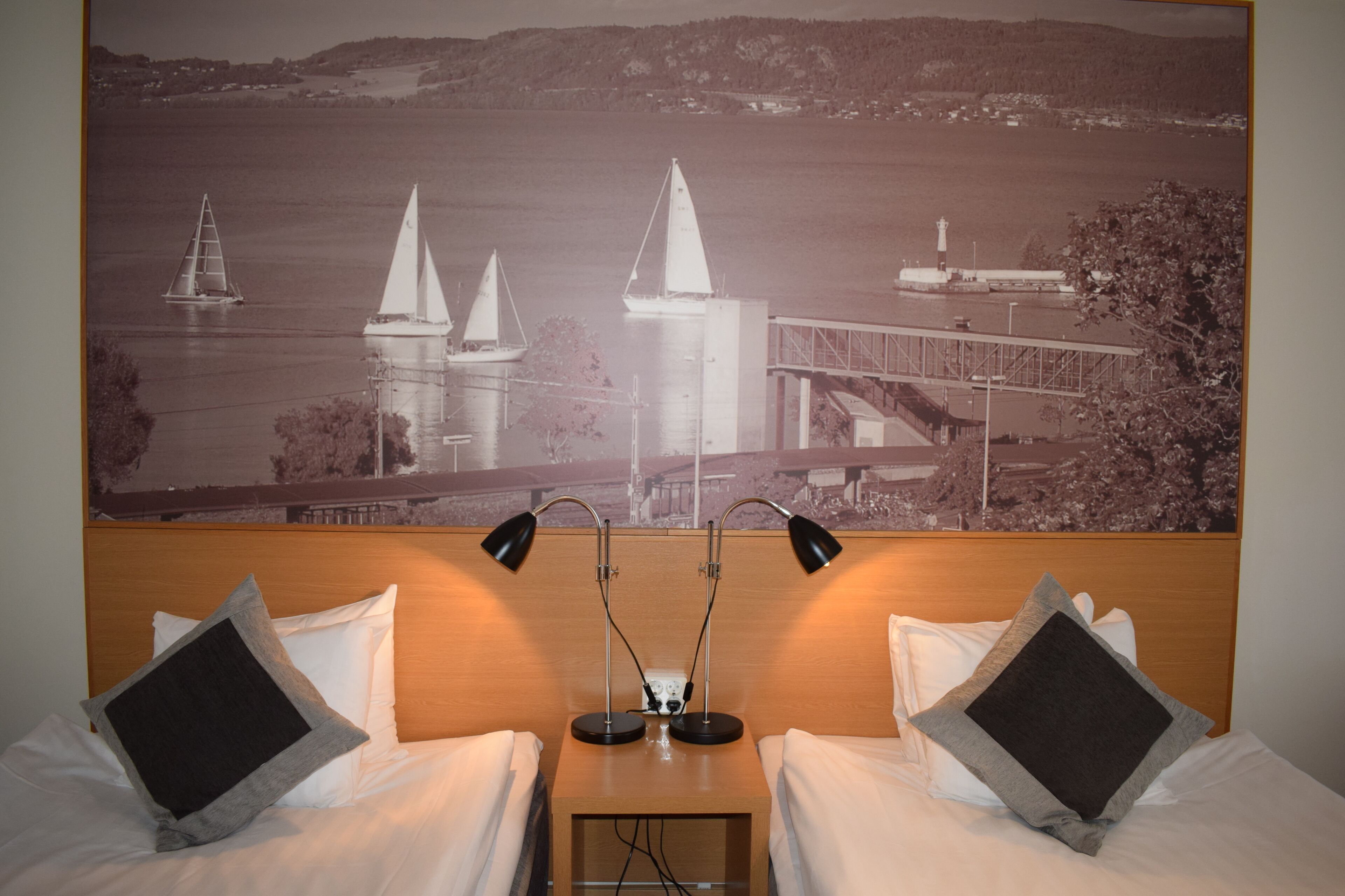 Ein Hotelzimmer mit zwei Betten, einem Nachttisch mit zwei Lampen und einem großen Schwarzweißfoto einer Brücke und Segelbooten an der Wand.