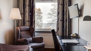 A room with a view of trees through a window, a desk with a computer, a chair, a lamp, and a radiator.