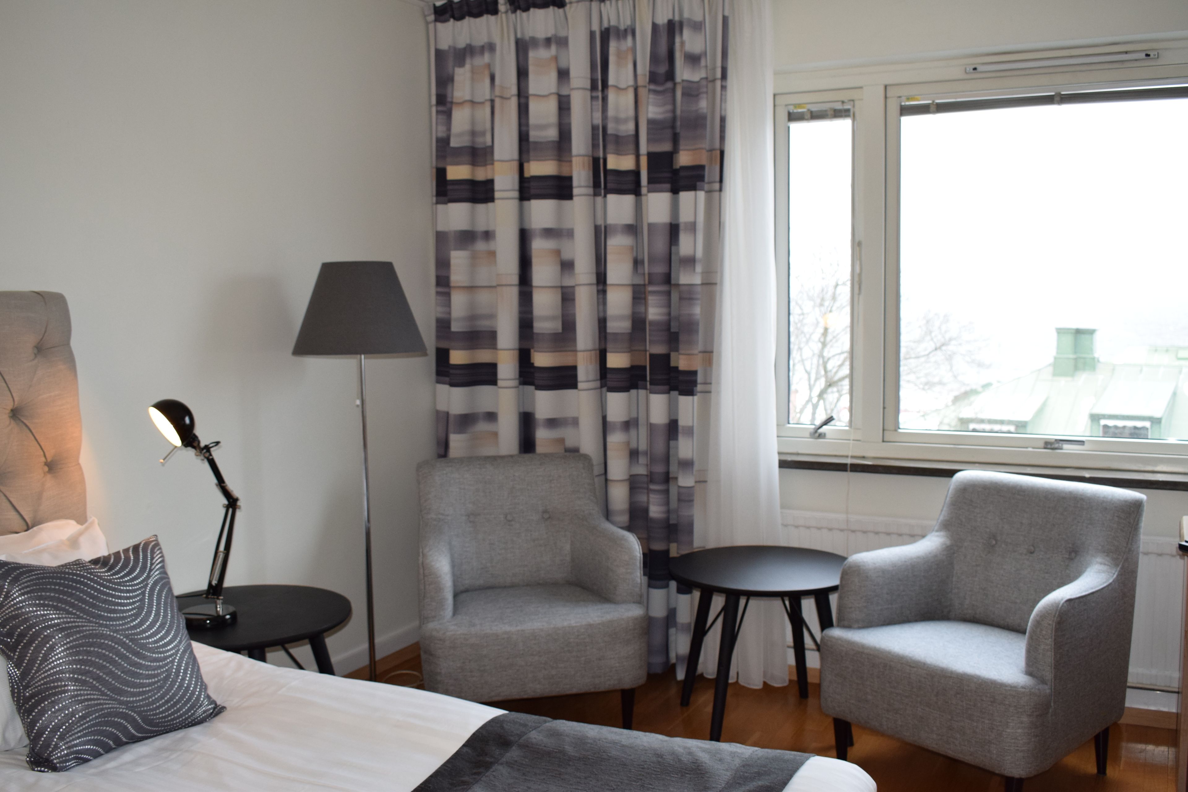 Habitación de hotel con una cama, dos sillones, una mesita, una lámpara y vista a la ciudad a través de una ventana.