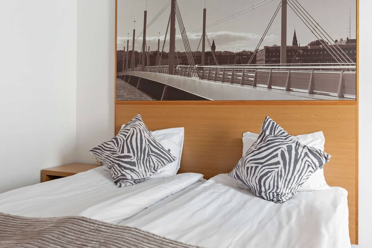 A hotel room with a bed featuring zebra-patterned pillows, a wooden headboard, and a view of a bridge with a cityscape in the background.