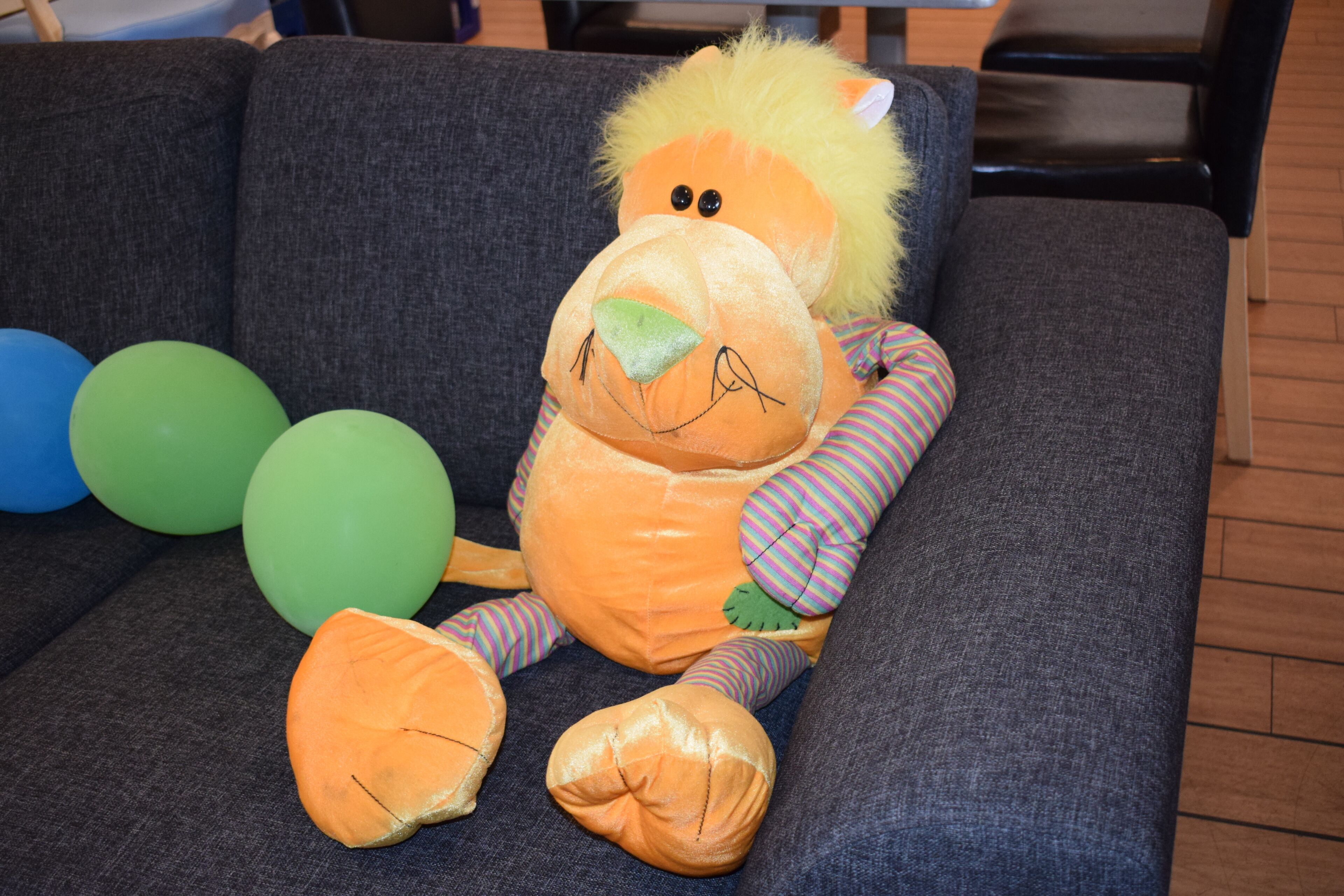 Un león de peluche sobre un sofá gris, cerca hay globos azules y verdes.