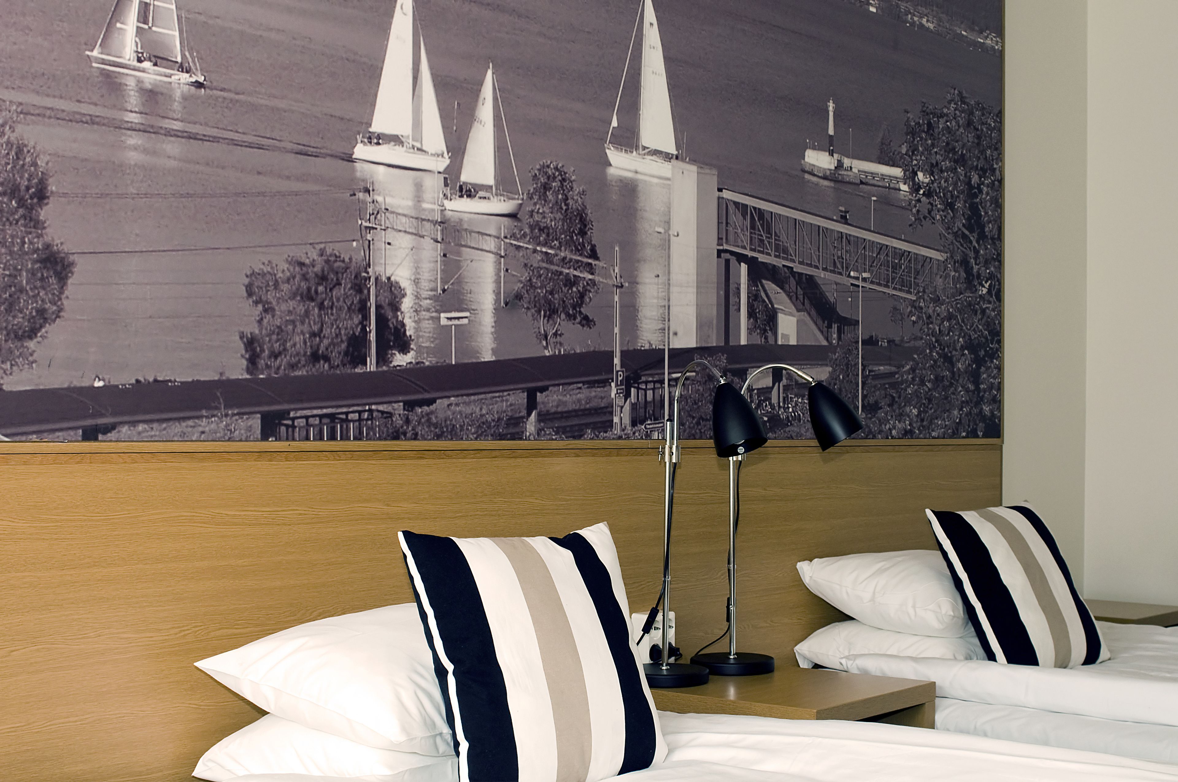 Habitación de hotel con cama, dos almohadas, mesita de noche, lámpara y un gran mural en la pared con barcos a vela y un puente.
