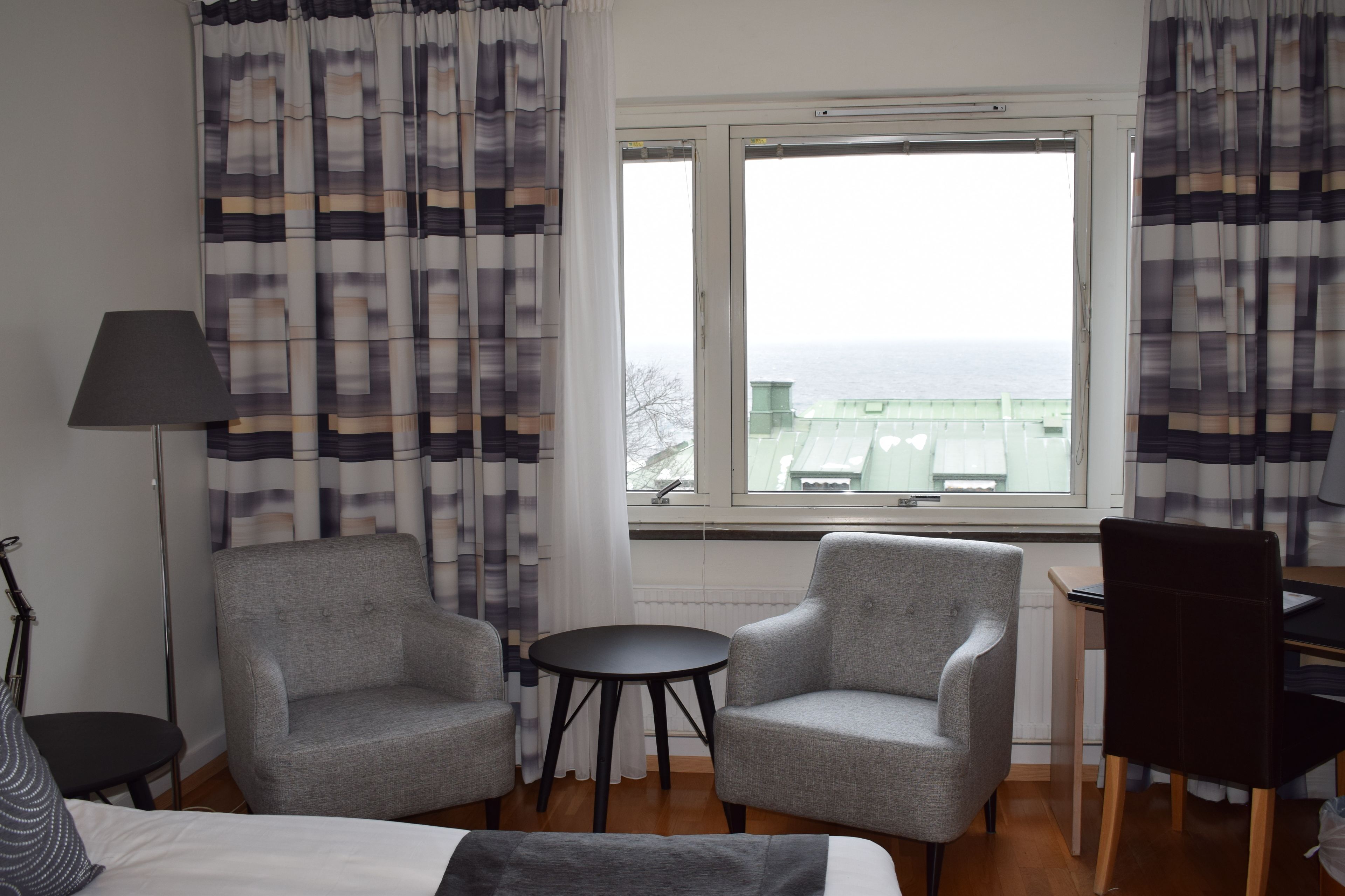 A hotel room with two grey armchairs, a small round table, a bed, a desk, and a view of the sea through a window.