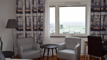 A hotel room with two grey armchairs, a small round table, a bed, a desk, and a view of the sea through a window.