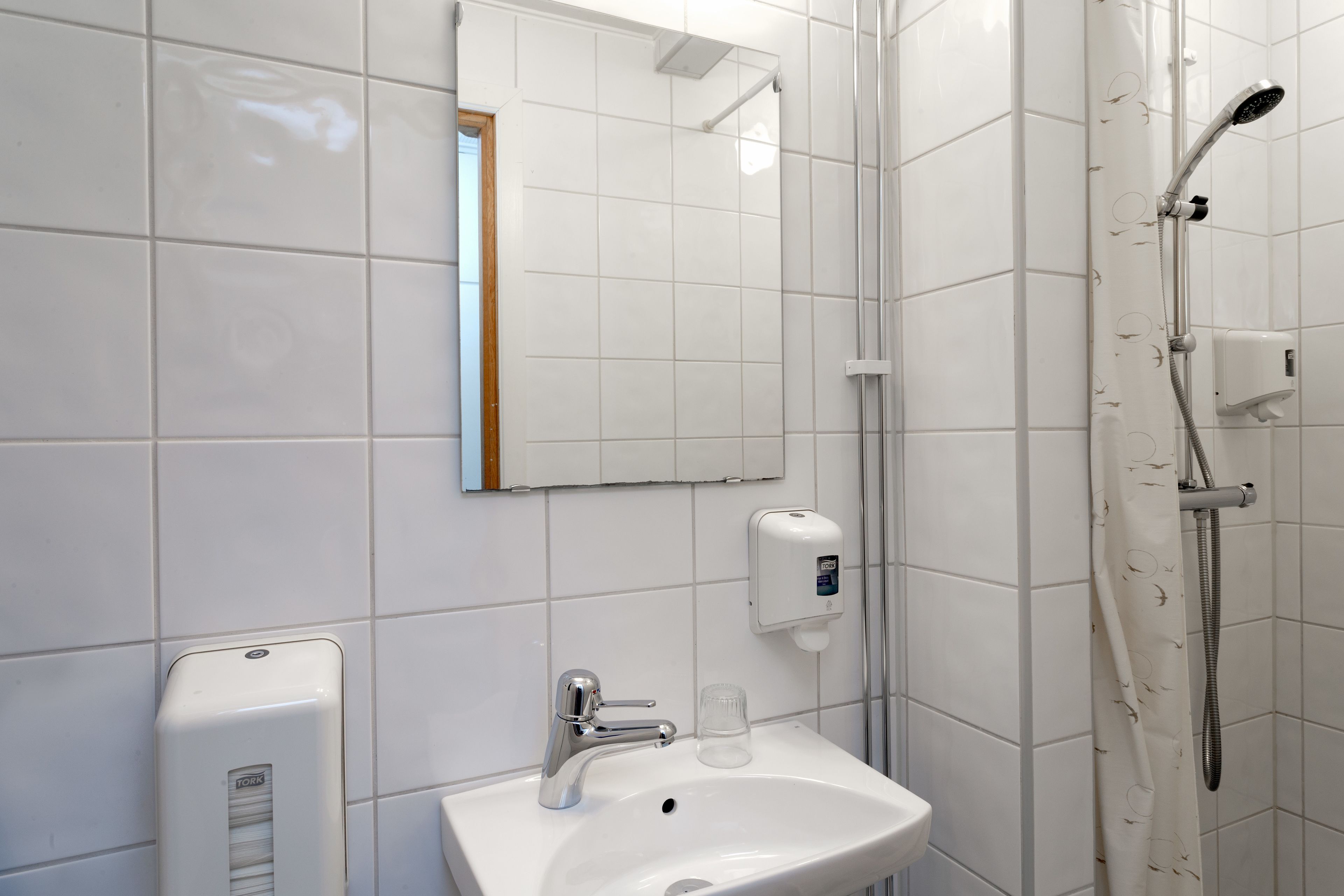 Un baño con azulejos blancos, un lavamanos con dispensador de jabón, un espejo y una ducha con cabezal de ducha manual.