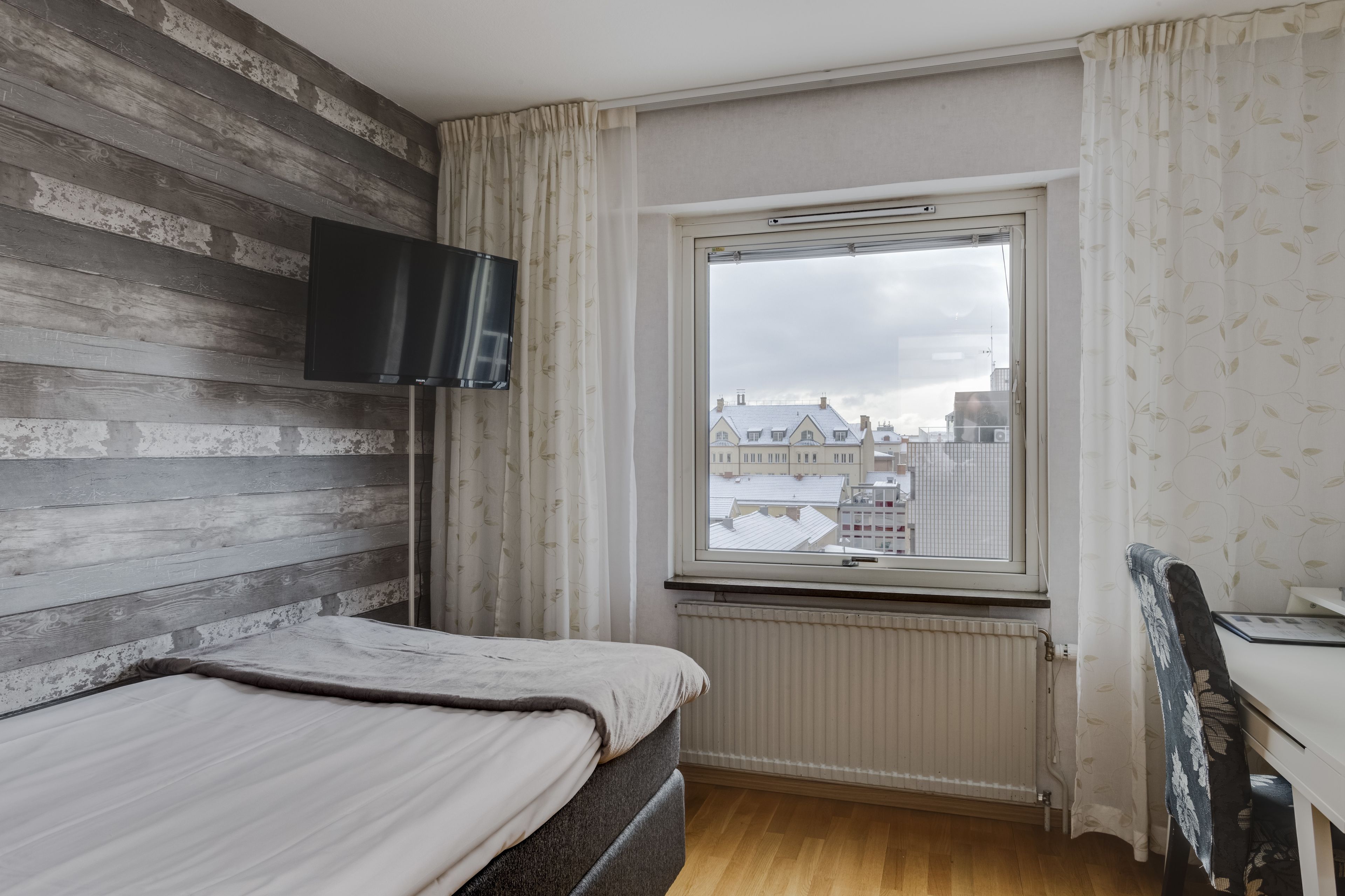 A bedroom with a bed, a TV mounted on the wall, a window with a view of buildings, and a radiator.
