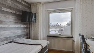 A bedroom with a bed, a TV mounted on the wall, a window with a view of buildings, and a radiator.