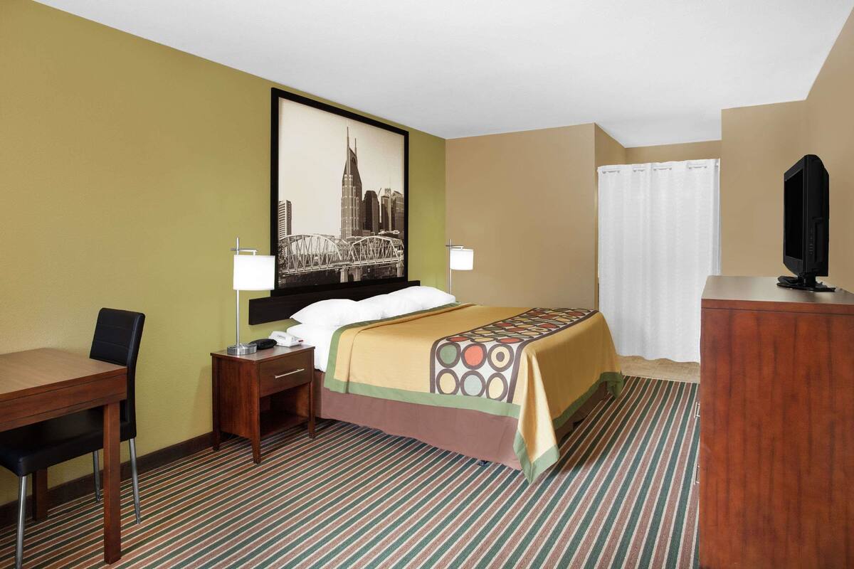 Ein Hotelzimmer mit einem Bett, einem Schreibtisch, einem Sessel, einem Fernseher und einem großen Stadtbild an der Wand.