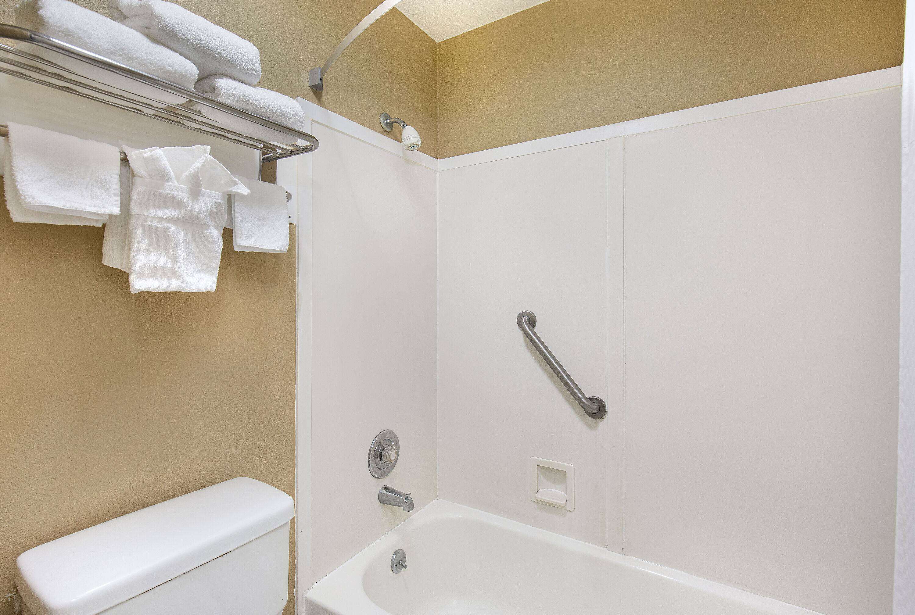 A bathroom with a white bathtub, a toilet, a towel rack with folded towels, and a handrail.