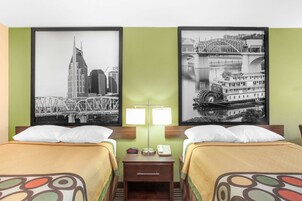 A hotel room with two beds, a nightstand with a lamp, and two framed cityscape pictures on the wall.
