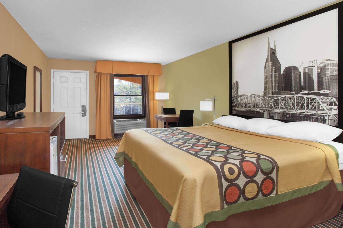 Ein Hotelzimmer mit Bett, Schreibtisch, Fernseher, einem Fenster mit Vorhängen und einem Stadtbildgemälde an der Wand.