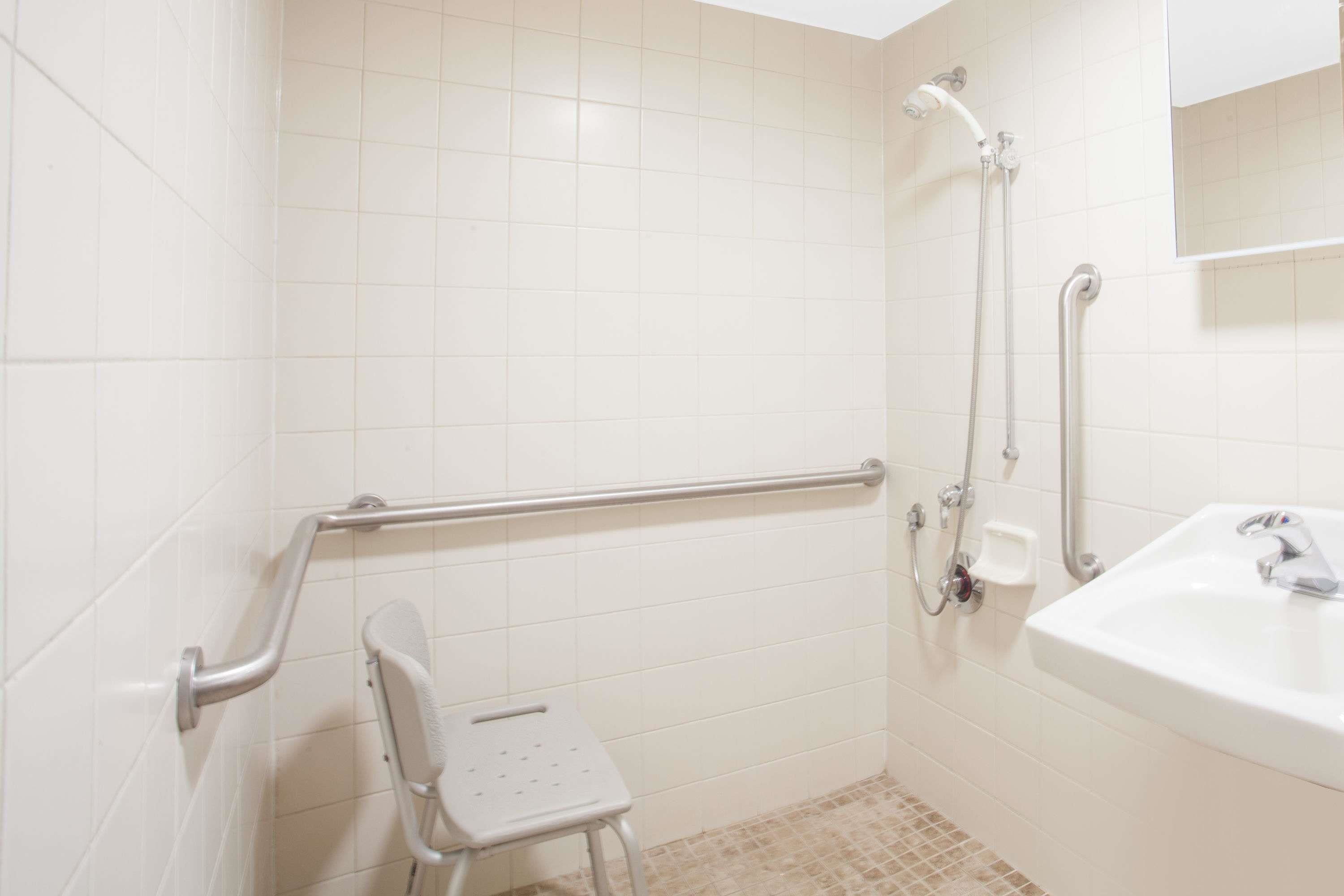 A bathroom with white tiles, a sink, a shower with a handheld showerhead, a grab bar, and a small stool.