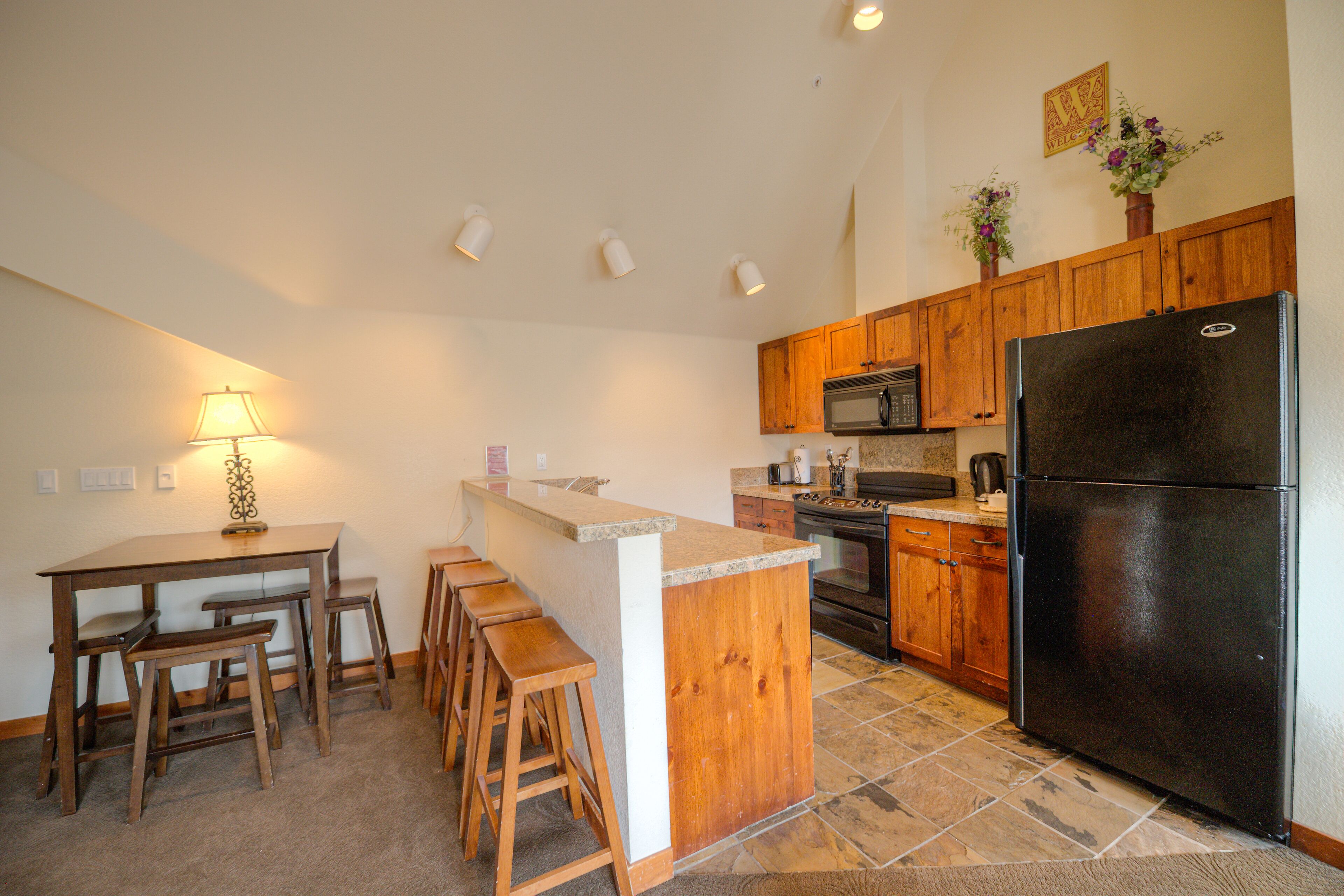 A kitchen with wooden cabinets, a black refrigerator, a microwave, and a bar area with stools.