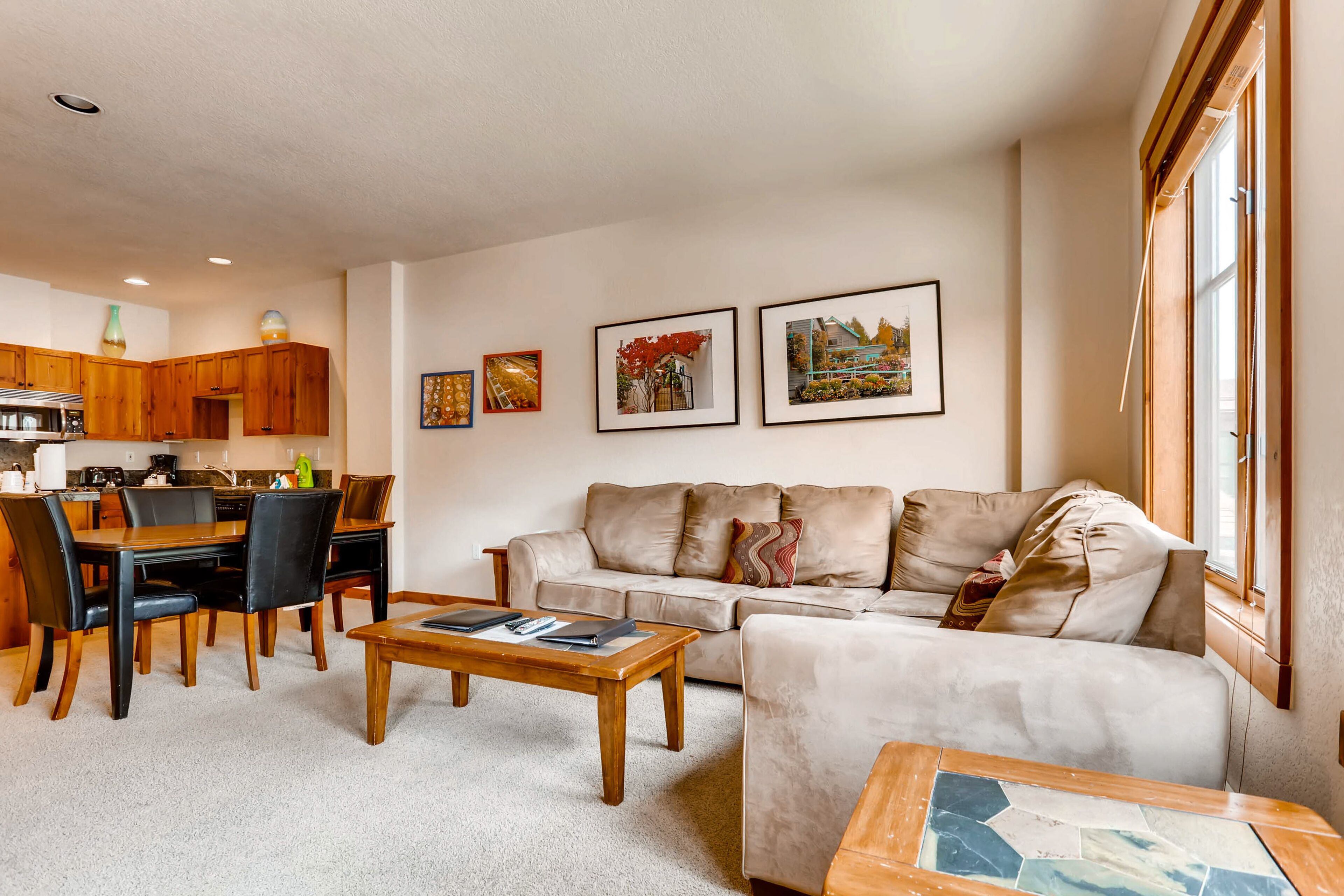 A living room with a beige sofa, a wooden coffee table, and a dining area with a wooden table and chairs. There are framed pictures on the wall and a window with blinds.