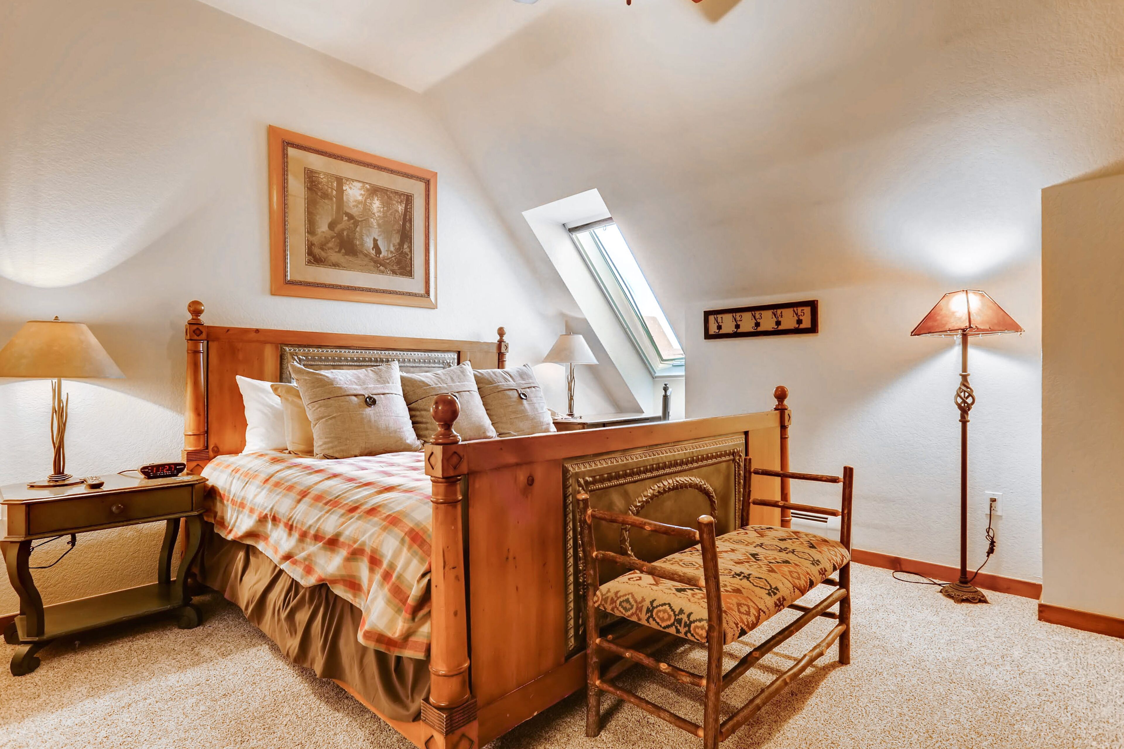 A bedroom with a wooden bed, a chair, a nightstand, a lamp, a painting, and a skylight.