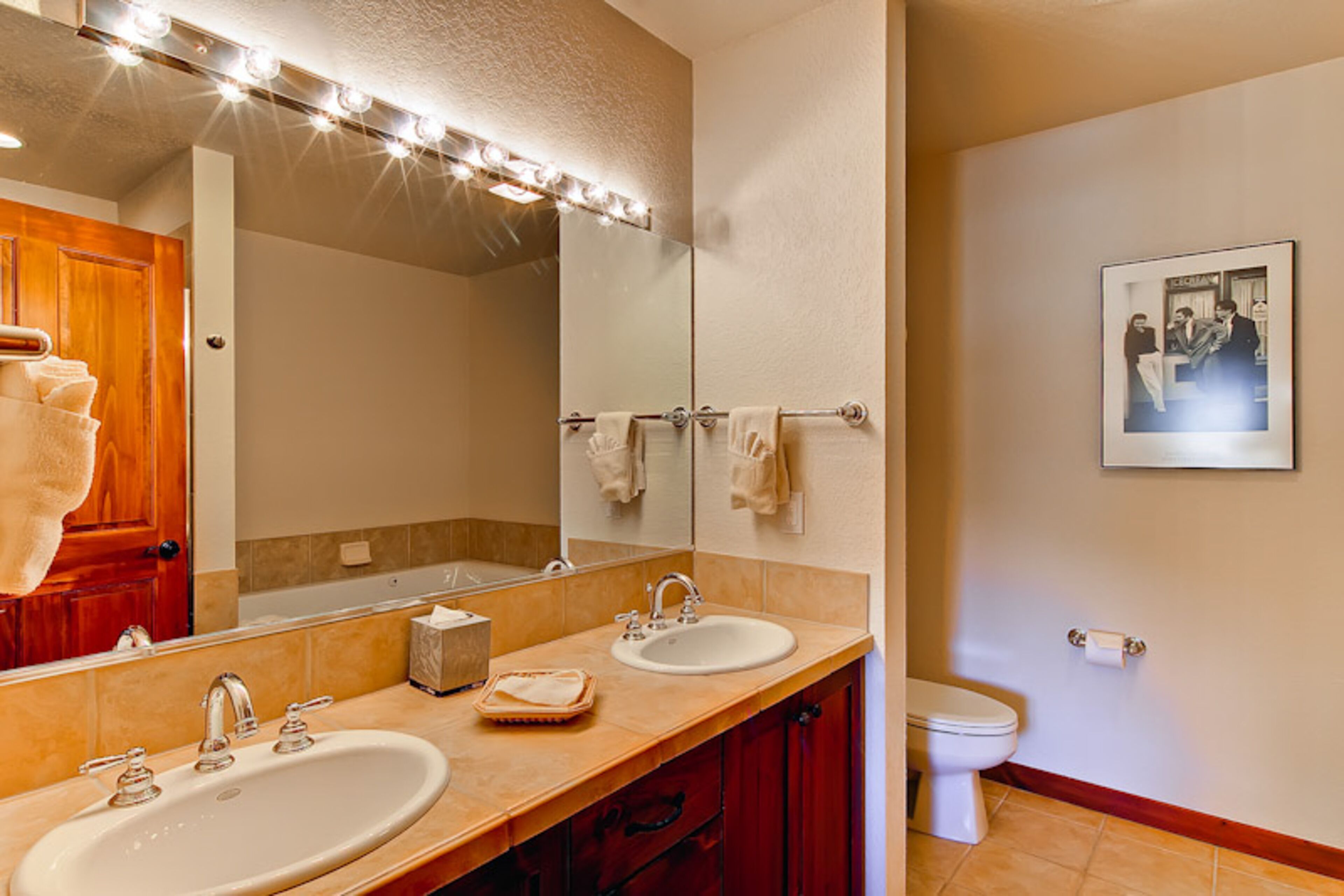 A bathroom with two sinks, a large mirror with lights, a towel rack, and a toilet.
