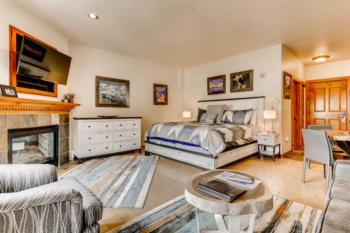 A bedroom with a bed, dresser, TV, fireplace, and a dining area with chairs and a table.