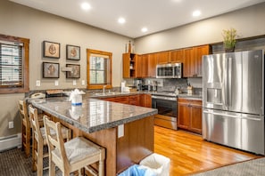 A spacious kitchen with wooden cabinets, a stainless steel refrigerator, granite countertops, and a central island with bar stools.