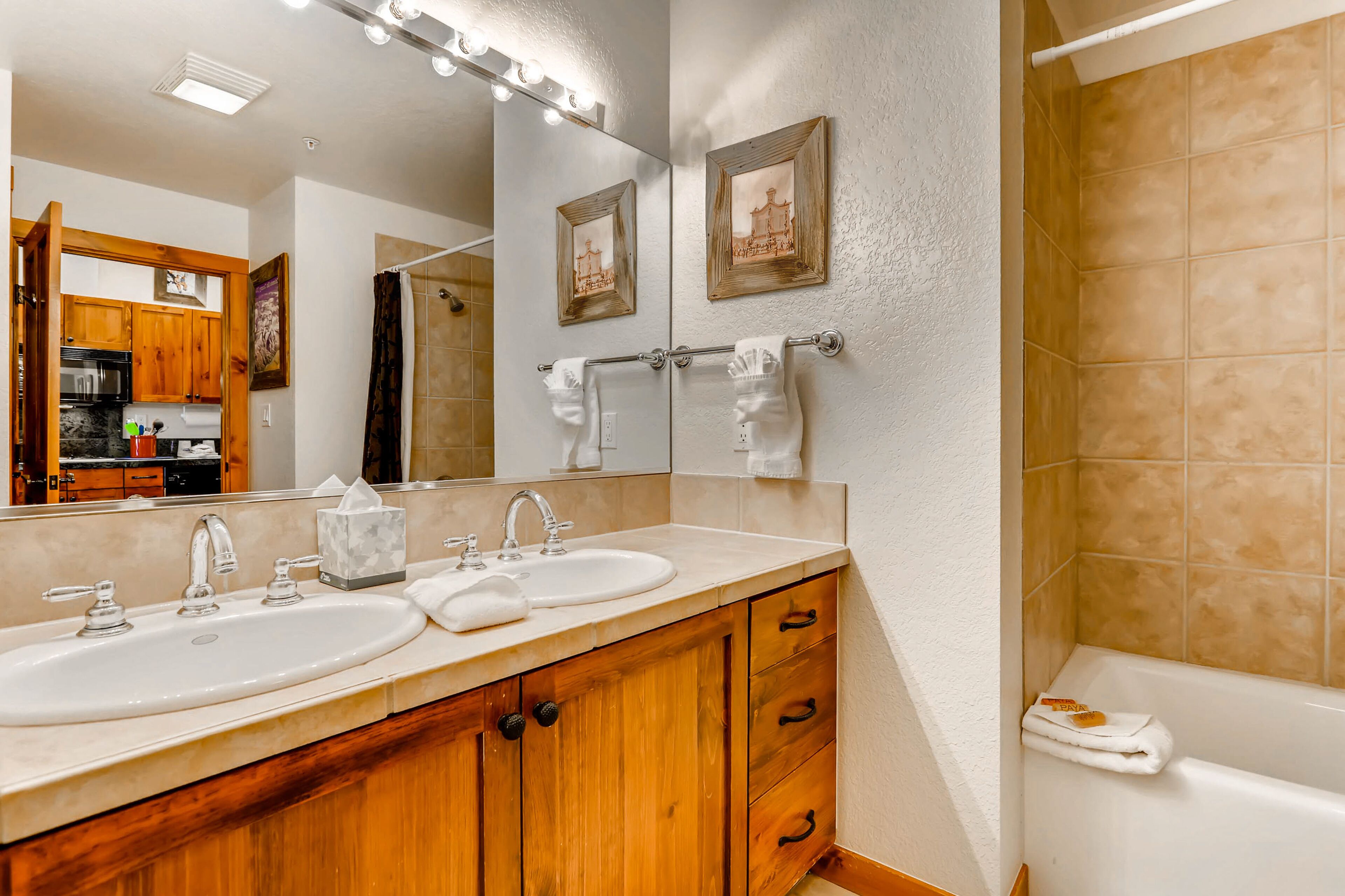A bathroom with two sinks, wooden cabinets, a mirror with lights, and a bathtub.
