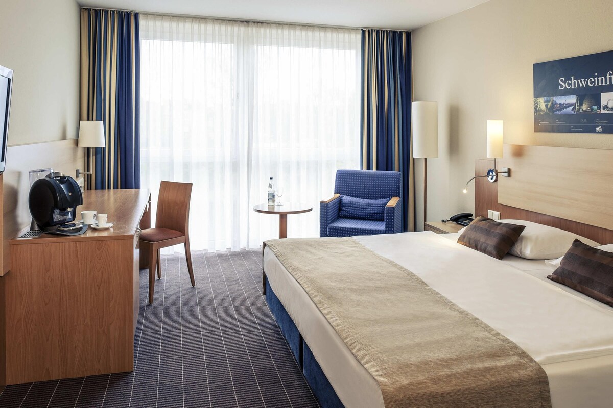 A hotel room with a bed, a desk with a coffee maker, a chair, a small table with a bottle and glass, and a lamp.