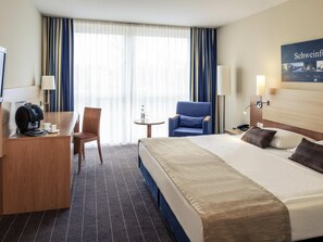 Une chambre d’hôtel comprenant un lit, un bureau avec une machine à café, une chaise, une petite table avec une bouteille et un verre, et une lampe.
