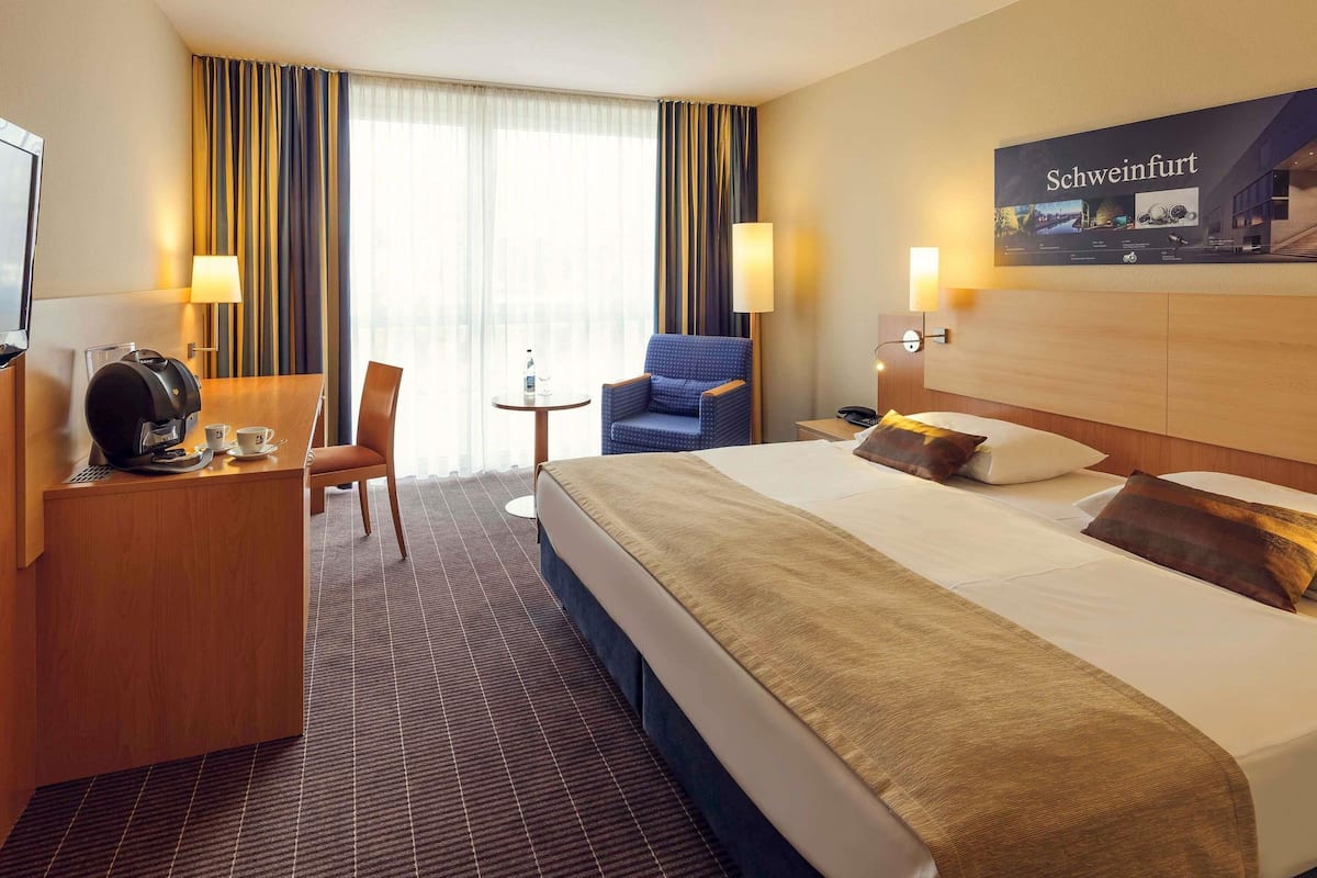 Een hotelkamer met een bed, een bureau met een koffiezetapparaat, een stoel, een klein tafeltje met een glas en een lamp.