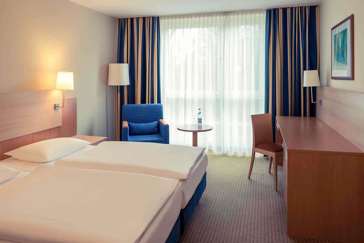 Een hotelkamer met twee bedden, een bureau, een stoel, een klein tafeltje en een groot raam met gordijnen.
