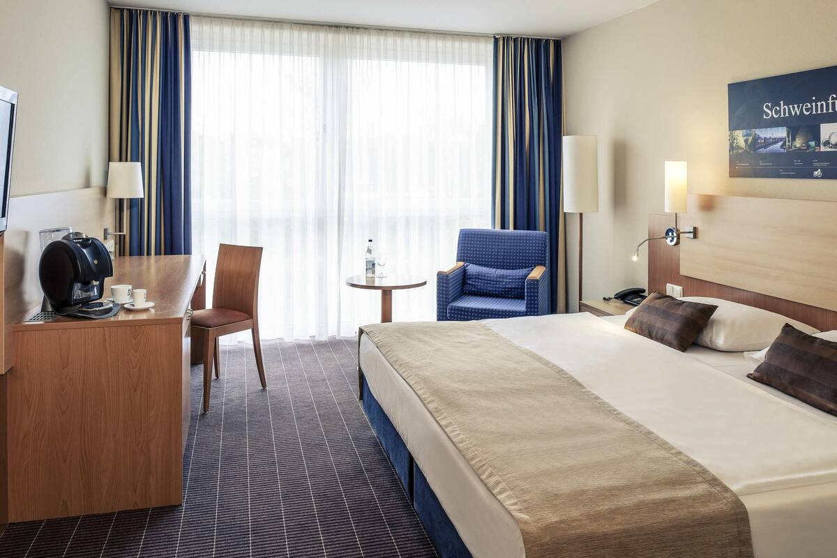 Een hotelkamer met een bed, een bureau met koffiezetapparaat, een stoel, een klein tafeltje met een fles en glas, en een lamp.