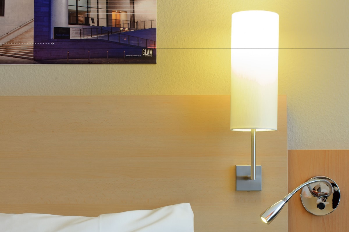 A hotel room with a bed, a wall-mounted lamp, a wooden headboard, and a view of the hotel's interior.