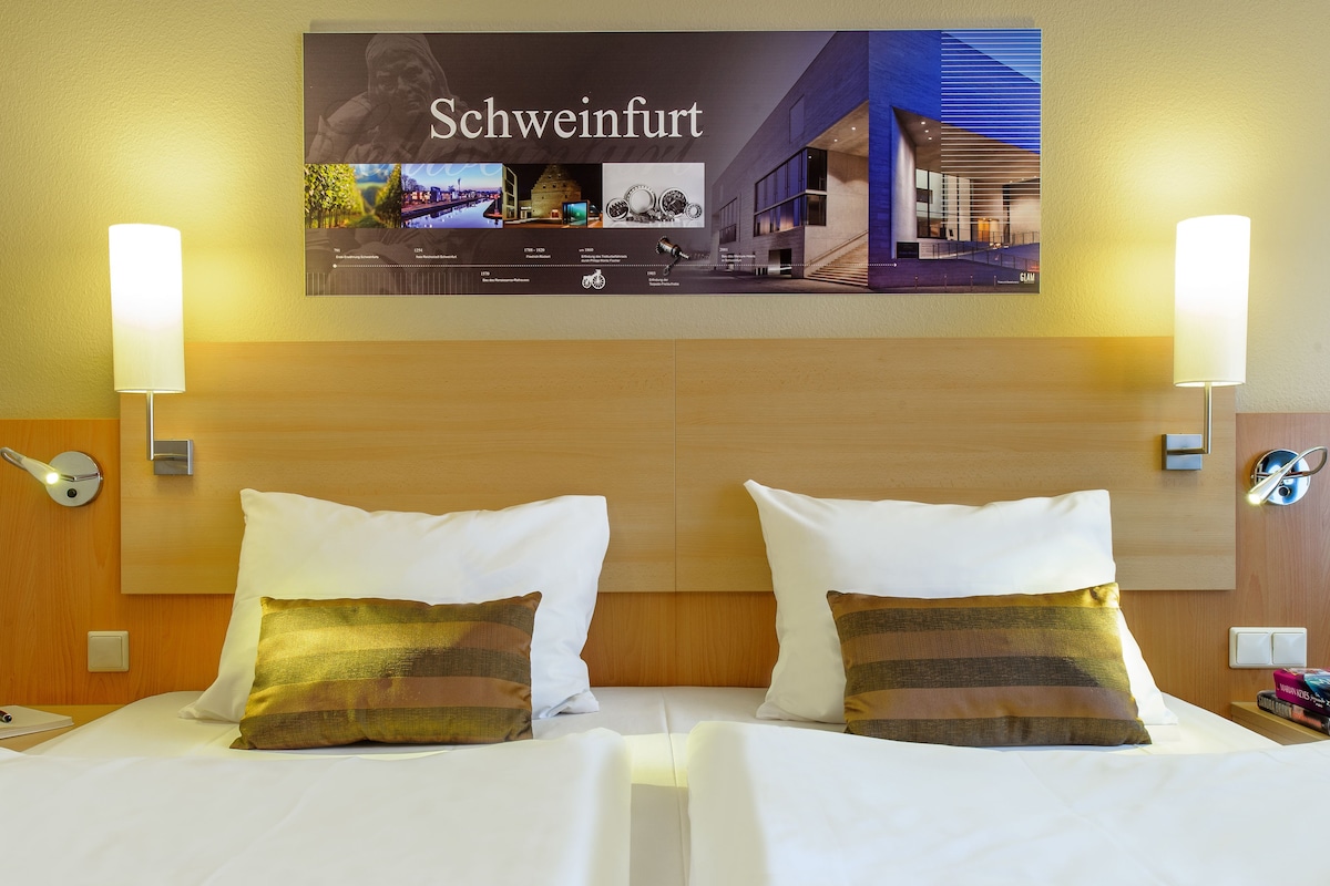 A hotel room with two beds, a wooden headboard, and a wall-mounted poster of Schweinfurt.
