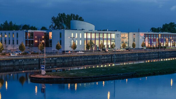 Un hôtel moderne doté d’une grande façade en verre, d’un plan d’eau devant et d’un parking.