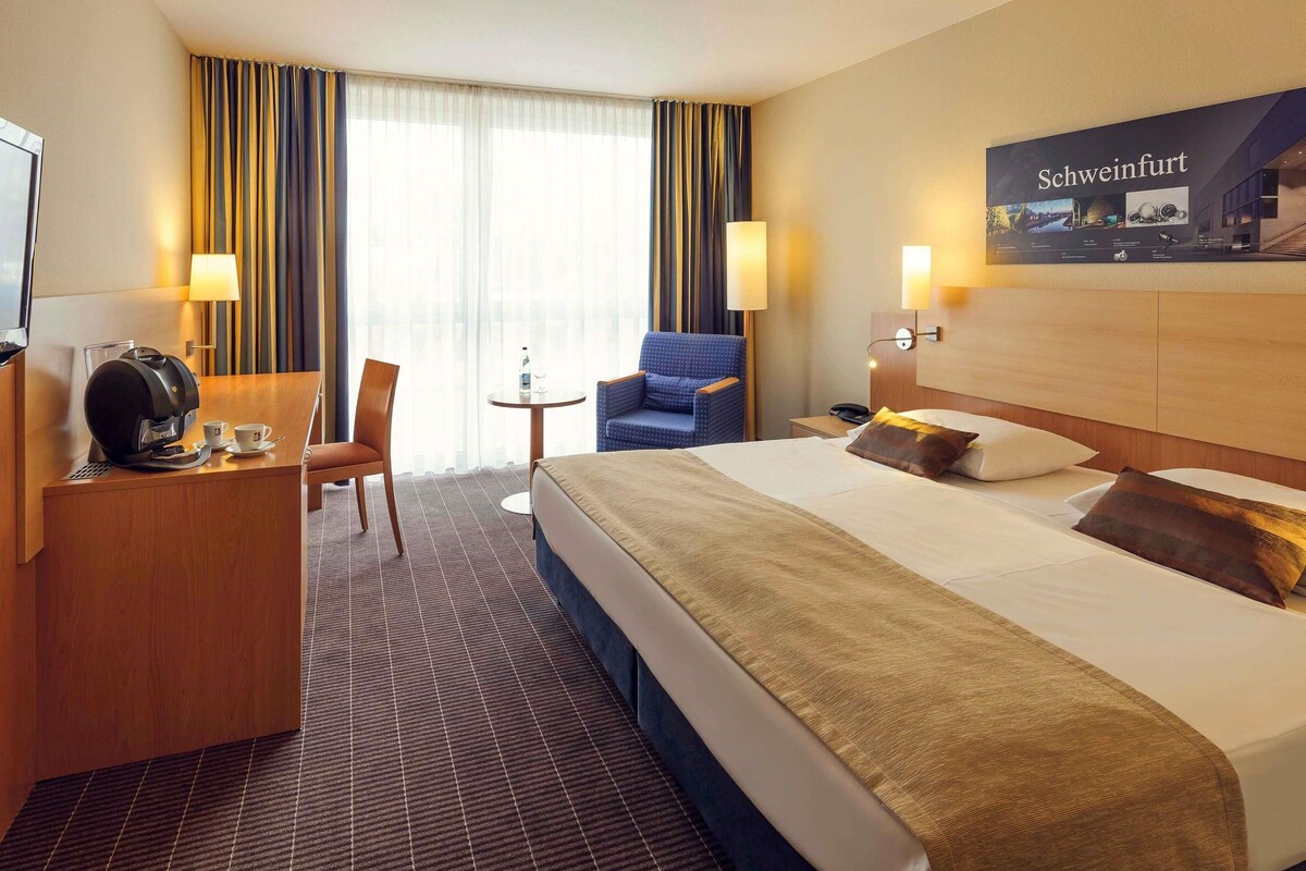 A hotel room with a bed, a desk with a coffee maker, a chair, a small table with a glass, and a lamp.